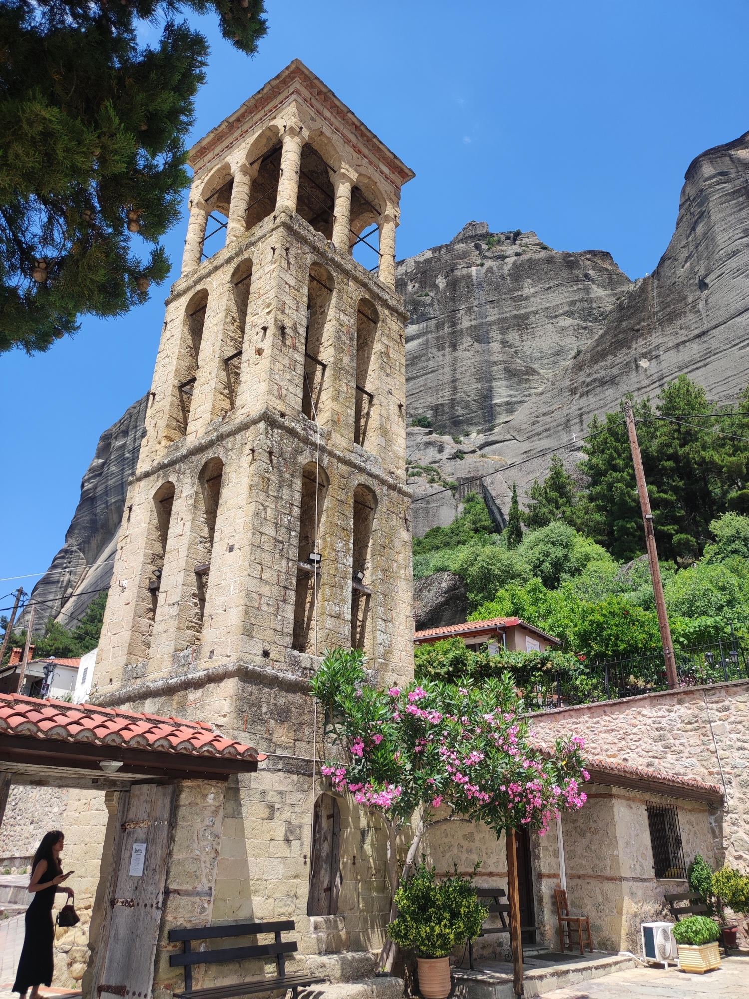 In Kalabaka, Greece, a stone bell tower with arched openings stands prominently in the foreground. To the left of the tower, a woman in a black dress stands near a wooden doorway, holding a handbag. A pink flowering tree is situated in front of a stone building with a tiled roof. In the background, towering rock formations rise above lush green trees, with a clear blue sky overhead. The scene captures a peaceful outdoor setting with architectural and natural elements.