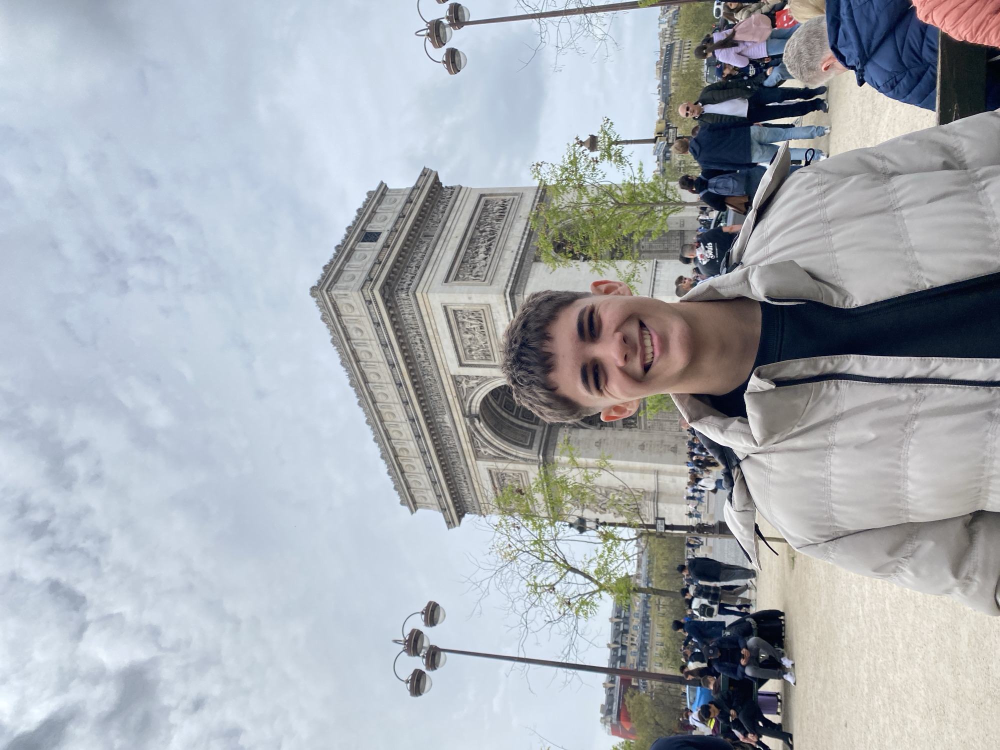 A young man smiles at the camera in front of the Arc de Triomphe in Paris, France. He is wearing a light-colored puffy jacket over a dark shirt. Behind him is the Arc de Triomphe, with architectural details visible. People are walking around the monument. Lamp posts are visible, and trees with green leaves are scattered throughout. The sky is cloudy.