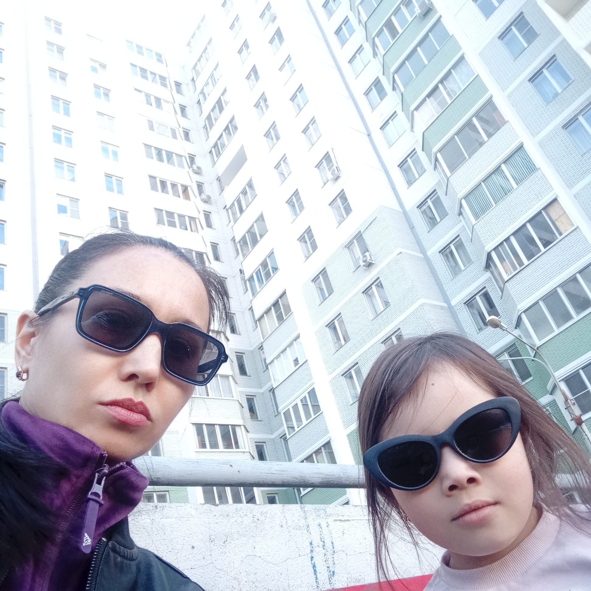 The image is a selfie of two individuals, presumably a mother and daughter, standing outdoors. The mother is on the left side of the frame and is wearing dark sunglasses and a jacket with a purple collar. The daughter is on the right, also wearing sunglasses and a light-colored shirt. Behind them is a tall building with numerous windows, giving the impression of an urban environment. The building is characterized by a light color with alternating horizontal sections of white and a bluish-green hue. The background is slightly overexposed. The location is Izhevsk, Russia, as indicated by architectural style and local knowledge.
