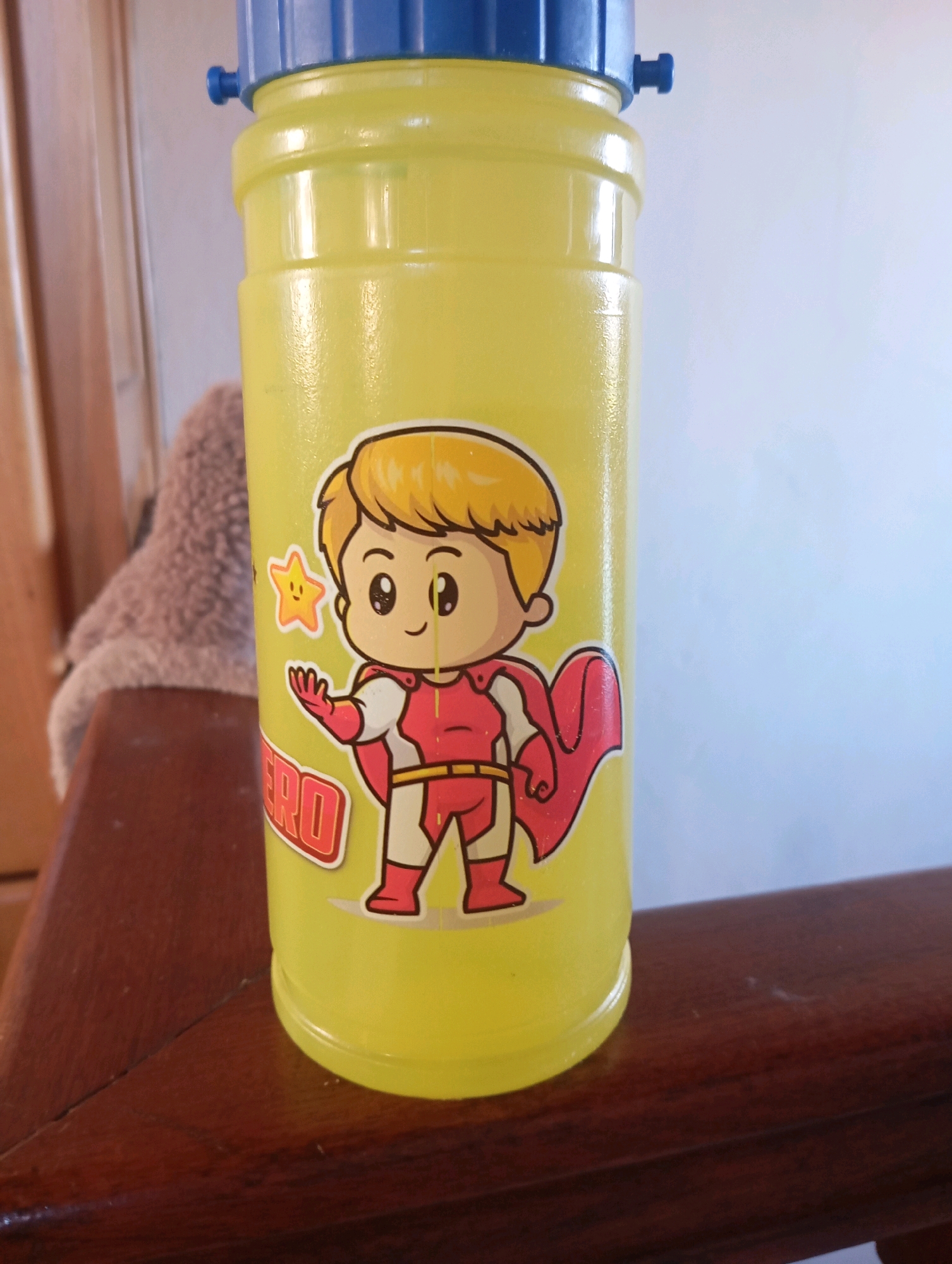 A bright yellow plastic bottle with a blue cap sits on a wooden surface. The bottle features a cartoon sticker of a child dressed as a superhero, complete with a red cape and costume, and a small yellow star. The word "HERO" is visible on the bottle below the sticker. In the background, a blurred image of a tan stuffed animal and wooden furniture can be seen, along with a pale wall. There is no indication that this image was taken in Viseu, Portugal.