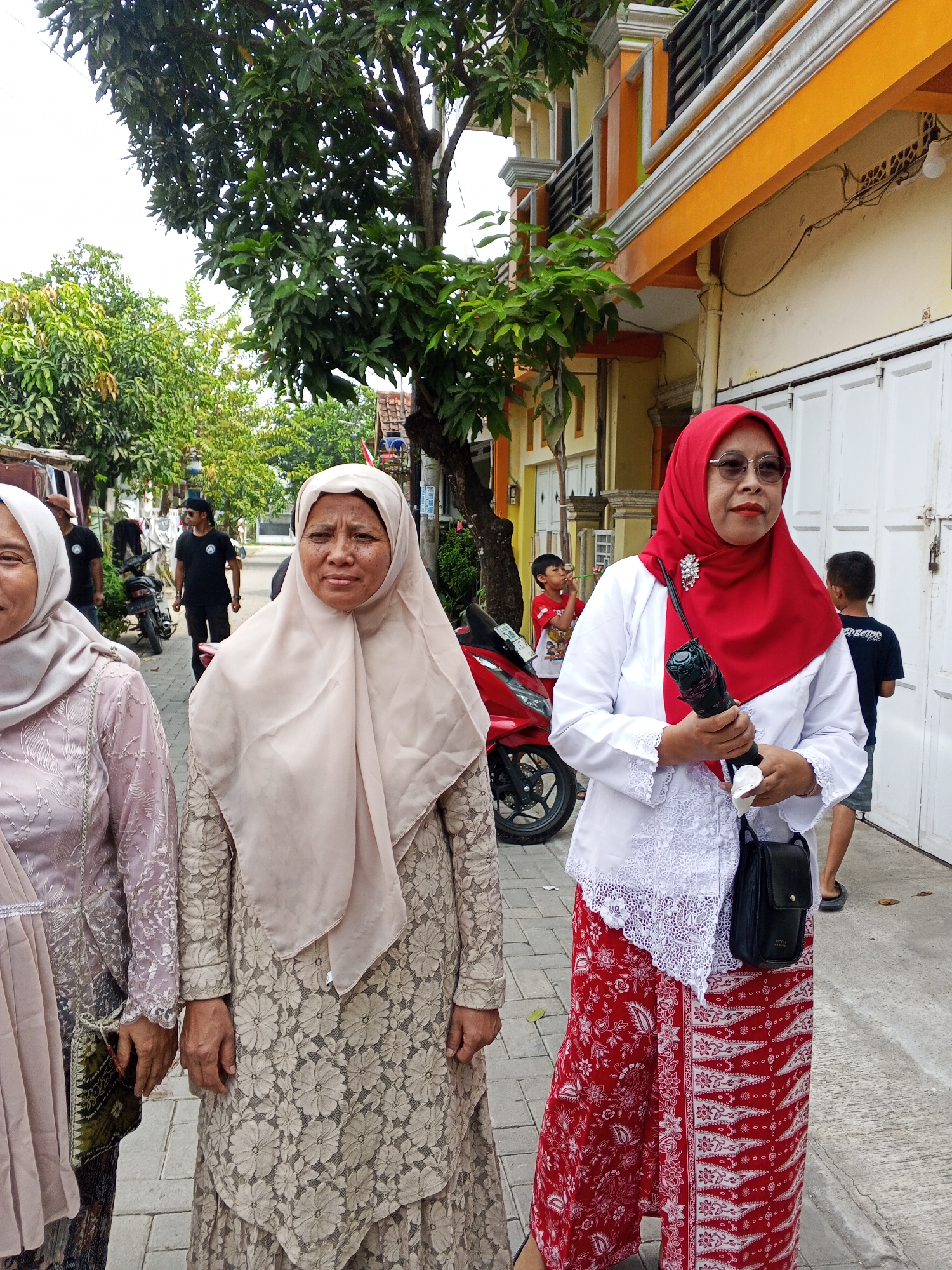 In the city of Ciruas, Indonesia, three women are standing on a paved sidewalk in front of a building. The woman on the right wears a red hijab, a white long-sleeved shirt with lace trim, and red pants with a white floral pattern. She is holding a black object, possibly a walkie-talkie or a phone, in her right hand and a white paper in her left hand. She also has a black bag slung over her shoulder. She is wearing sunglasses and a brooch on her shirt. The woman in the middle wears a beige hijab and a beige lace dress. The woman on the left wears a light pink hijab and a matching light pink dress with lace details. Behind the women, a red motorcycle is parked. Further in the background, a few men and children are visible, along with trees and a building with orange and yellow accents. The overall scene appears to be a street in a residential area.