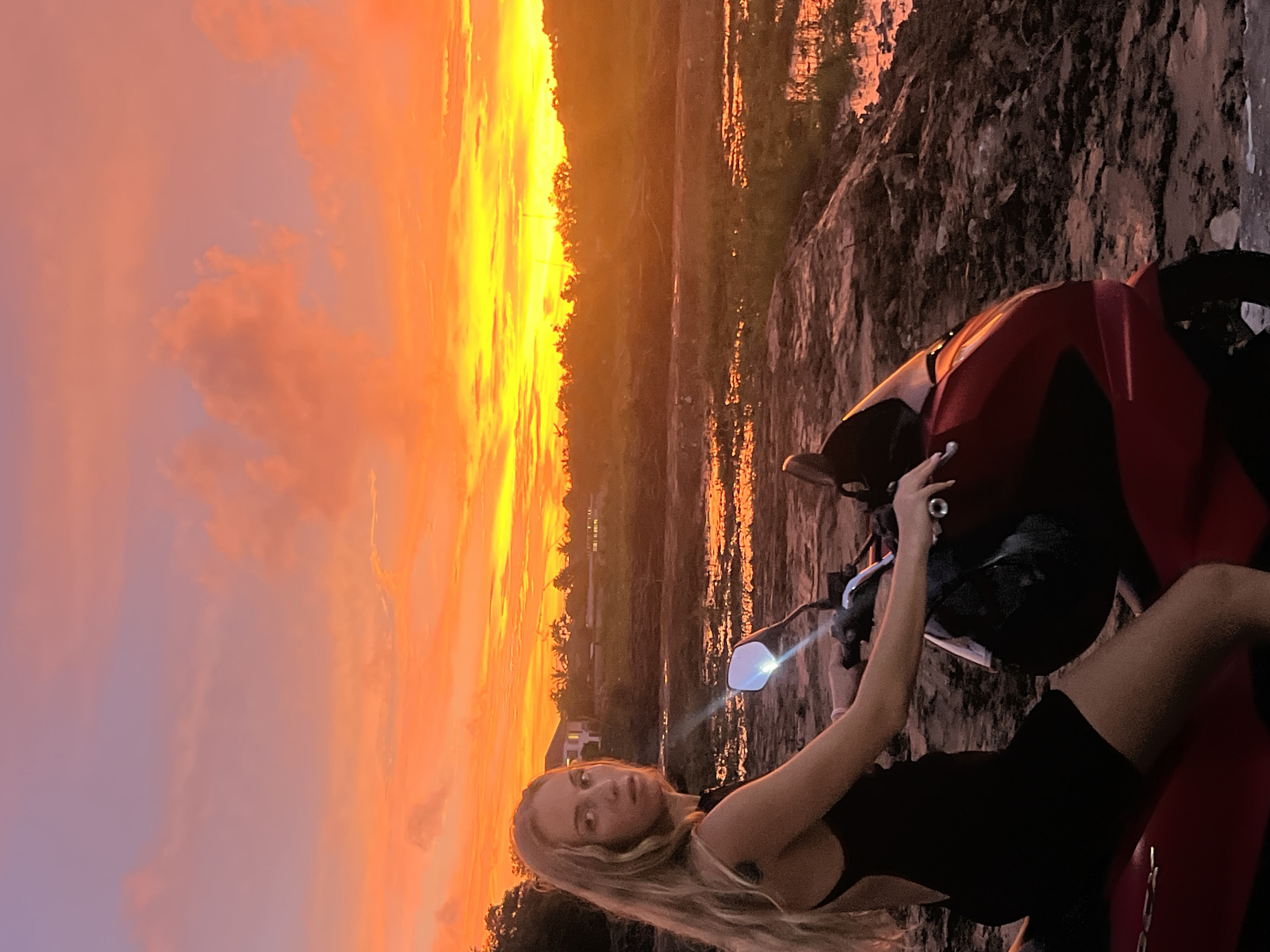 A young woman with long blonde hair, wearing a black dress, is seated on a red scooter. She is looking towards the camera with a neutral expression. The scooter is positioned on a muddy, uneven surface. Behind her, the sky is ablaze with a vibrant sunset, featuring shades of orange, yellow, and pink. In the distance, silhouetted trees and buildings can be seen. The setting appears to be a rural area outside of Thep Krasatti, Thailand, possibly near a body of water or wet land, given the muddy ground. The lighting suggests it is either sunrise or sunset.