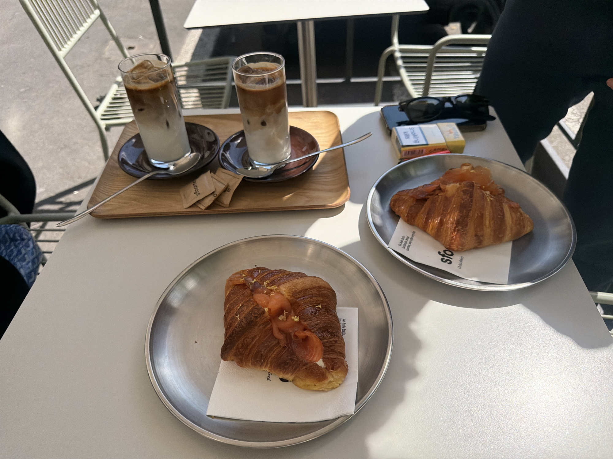The image shows a table at a cafe, likely in Milan, Italy. The table is occupied by two iced coffees on a wooden tray with brown sugar packets and spoons, two plates each containing a croissant topped with what appears to be smoked salmon, a pack of cigarettes, and sunglasses. A person is seated at the table, partly visible on the right side of the frame. The environment suggests an outdoor cafe setting.