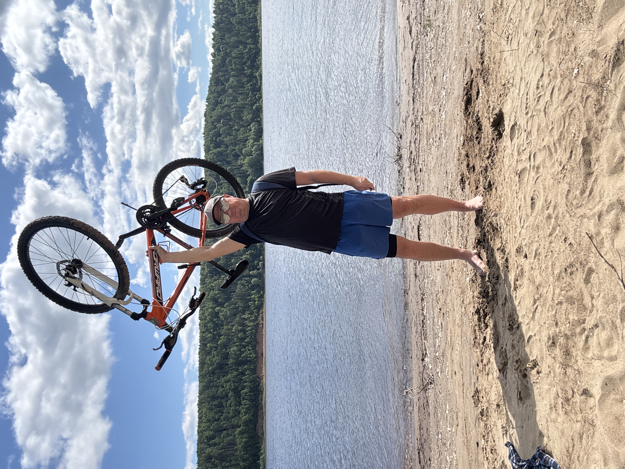 A man stands on a sandy beach, holding an orange mountain bike upright. He is wearing a black t-shirt, blue shorts, and a grey baseball cap. Sunglasses are perched on his nose. The man is looking towards the camera and smiling. The bike is partially obscuring him from the waist up.
The setting is a beach on a bright, sunny day. The water of a lake or river stretches out behind the man, with a dense forest lining the opposite shore. The sky above is a vibrant blue, dotted with fluffy white clouds. The sand appears somewhat damp near the water's edge, with footprints visible.
The image appears to be taken during daylight hours, likely late morning or afternoon, given the brightness and the shadows cast. The overall mood seems relaxed and outdoorsy, suggesting a recreational activity like cycling and a visit to the beach. There is no visible text in the image. The location is Ol'khovskoye, Russia.
