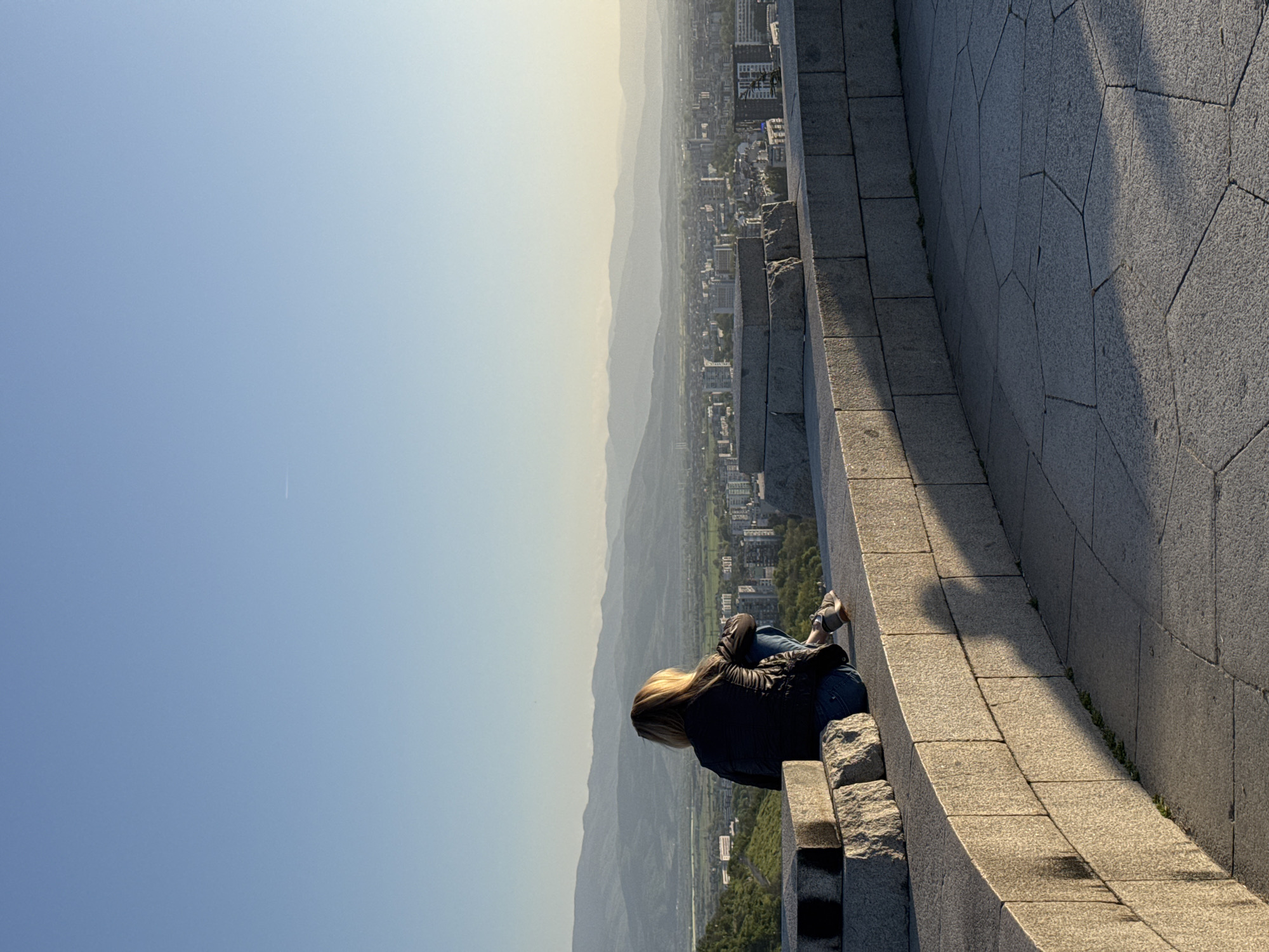 The image, when viewed in its correct landscape orientation, depicts a solitary person seated on a textured stone wall, gazing out over a vast panoramic view. The subject, seen from behind, appears to be a woman with long blonde hair, wearing a dark jacket and blue jeans. She is positioned on the right side of the frame, seemingly absorbed in the distant scenery.
The sturdy wall, constructed from grey stone blocks – a mix of rectangular and polygonal shapes – extends diagonally across the foreground and midground. Strong, elongated shadows are cast by the low-angle sunlight, indicating either early morning or late afternoon. Beyond the wall, a sprawling cityscape is visible in the mid-distance, nestled within a wide valley. Further in the background, multiple layers of hazy mountains or hills recede towards the horizon, creating a sense of atmospheric depth. The sky is clear, displaying a gradient from a light blue overhead to a softer, warmer hue closer to the horizon. A faint white contrail from an aircraft can be seen in the upper left sky.
The scene evokes a sense of peace and contemplation, with the individual enjoying the expansive view from a high vantage point, characteristic of elevated locations in cities such as Plovdiv, Bulgaria. No discernible text is visible in the image.