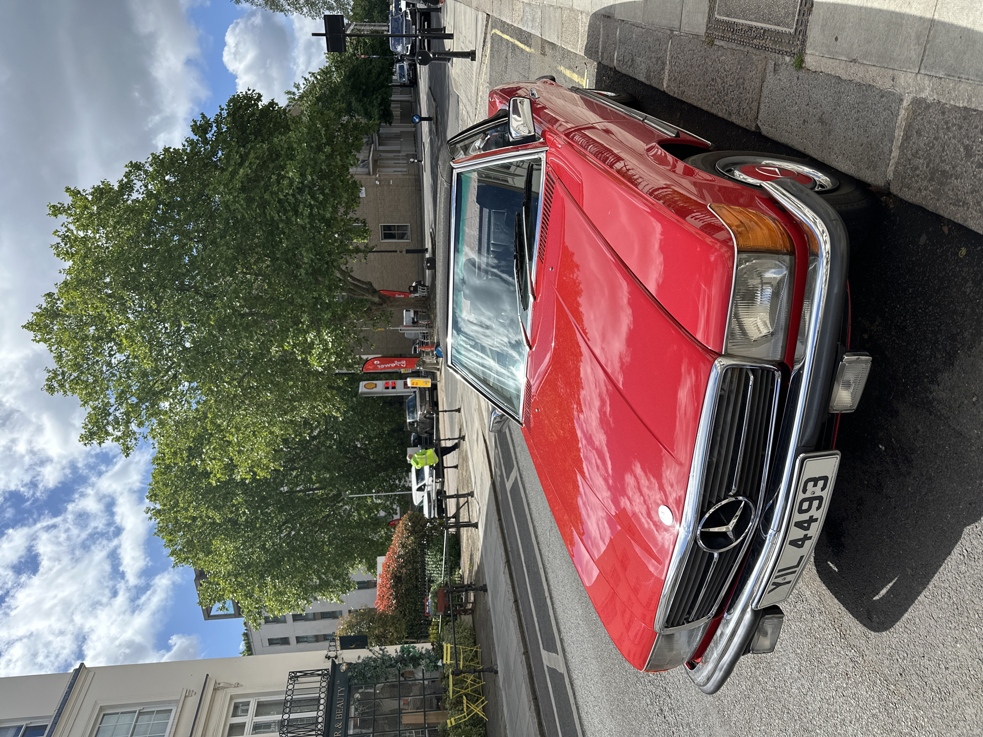 A red Mercedes-Benz SL is parked on a London street. The registration plate reads "YIL 4493". The car is facing right, with its front end close to the curb. A tree dominates the left side of the frame, with a building and street signs visible behind it. A few people are walking on the sidewalk. The sky is cloudy with patches of blue. The location appears to be a street with retail establishments.