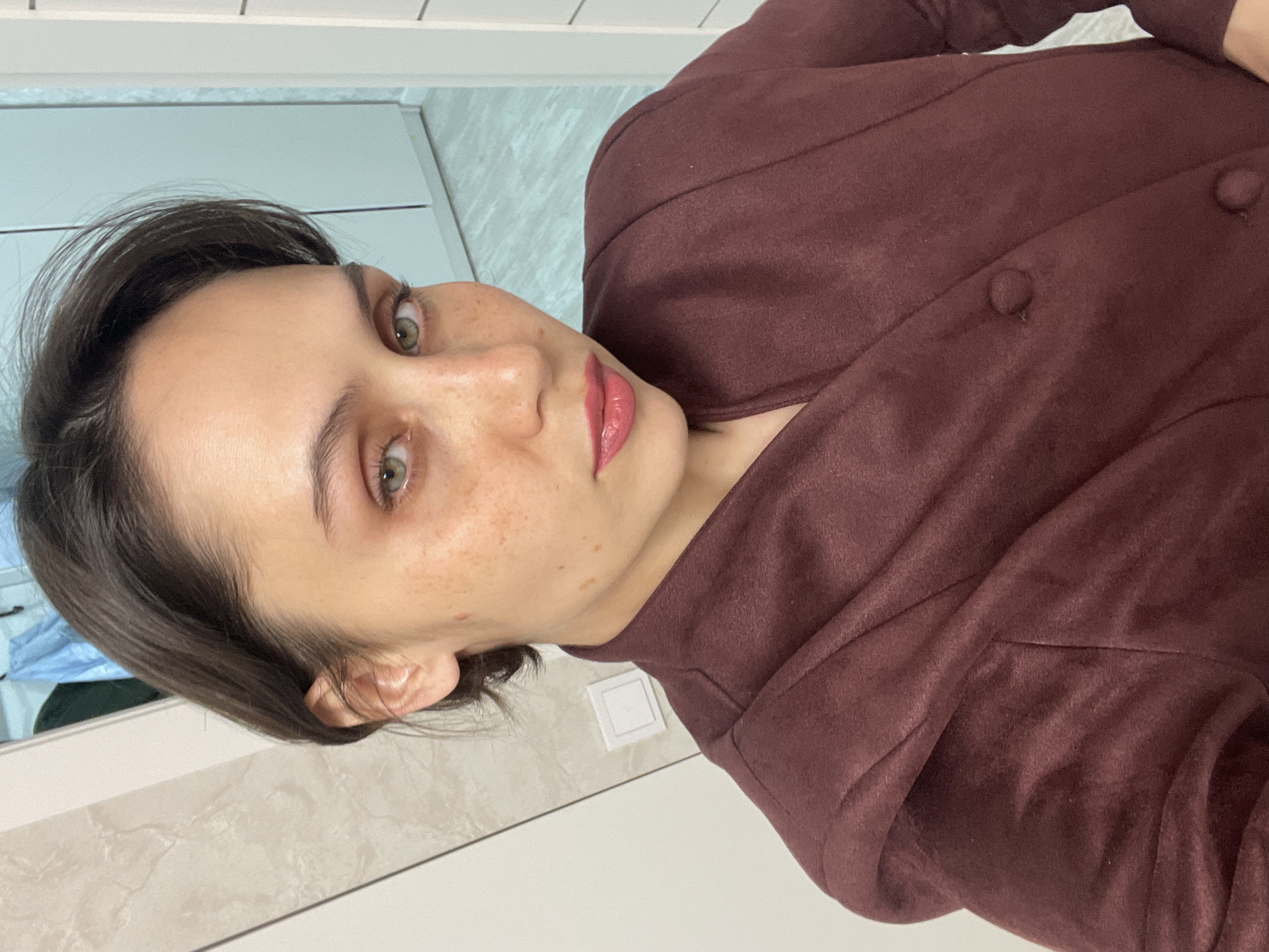 The image is an indoor, head-and-shoulders selfie featuring a young woman. She has short, dark brown hair and striking green eyes, which gaze directly at the camera. Her skin is light with numerous freckles visible across her nose and cheeks. She wears pink lipstick and has a neutral expression, possibly with a hint of thoughtfulness.
She is dressed in a dark reddish-brown, possibly plum-colored, jacket or coat made of what appears to be a soft, felt-like or suede material. Several buttons are visible on the front of her garment.
The background is an indoor setting, mostly out of focus, consisting of light-colored walls, possibly tiled, with horizontal lines. A white square object, likely a light switch or electrical outlet, is visible on the wall behind her right shoulder. The lighting suggests an artificial indoor light source.
The overall scene depicts a casual portrait taken in what appears to be a modern interior space, potentially in Izhevsk, Russia. There is no visible text in the image.