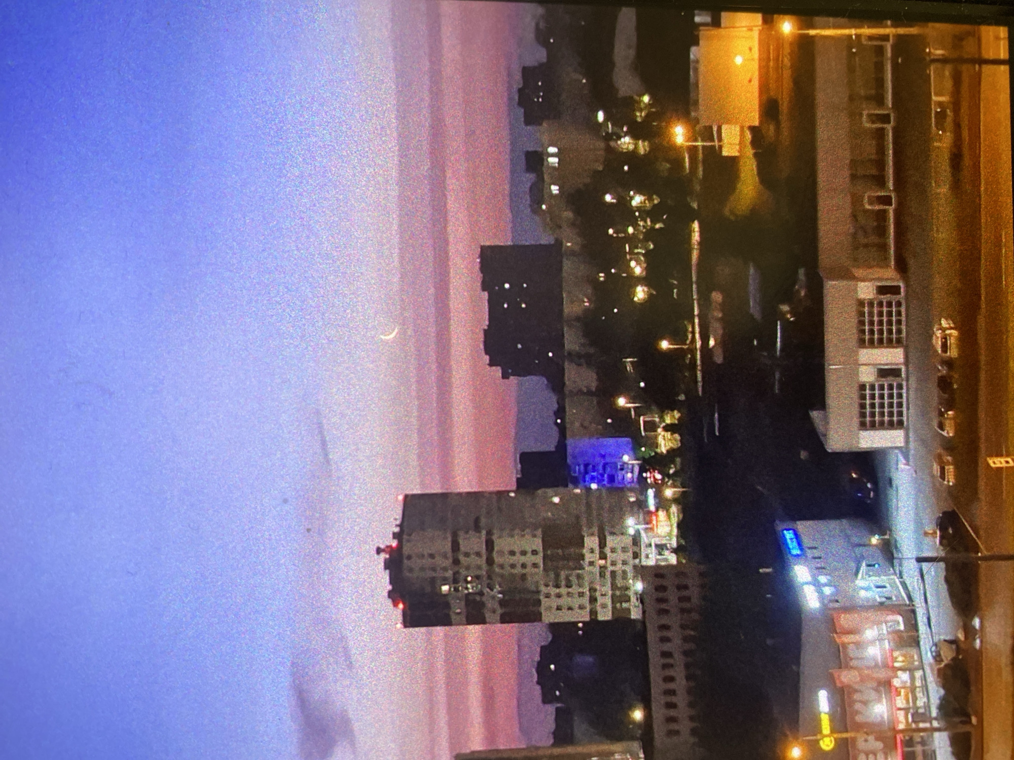 The media file, originally rotated counter-clockwise, depicts an urban landscape at twilight. The sky transitions gracefully from a deep blue at the top to soft pink and orange hues near the horizon, where a delicate crescent moon is distinctly visible.
The scene features numerous city buildings of varying heights and architectural styles, many with illuminated windows, suggesting active occupancy. A prominent multi-story building stands centrally, its facade and windows glowing with light. To the right, a street is lined with additional buildings, brightly lit by streetlights and commercial signs. These artificial lights cast warm yellow and orange tones, contrasting with the natural twilight. Several vehicles are subtly visible on the streets.
No specific human activity is evident, but the illuminated windows and active street lighting portray a bustling yet serene city as it transitions from day to night. A blurry blue sign on a building to the right appears to show Cyrillic text, consistent with the provided location context of Saint Petersburg, Russia. Another indistinct yellow and red commercial sign is also visible.
The image quality shows considerable digital noise or grain, particularly in the sky and darker areas, suggesting low-light photography or a capture from a screen. The overall impression is that of a calm and atmospheric urban evening or early morning.