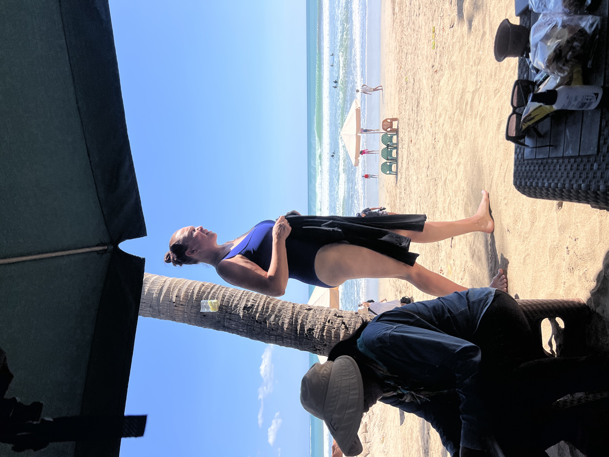 A medium shot captures a beach scene in Kuta, Indonesia, on a bright, sunny day. In the foreground, a person wearing a wide-brimmed hat and dark clothing is seated, looking down. To their right, a woman in a blue swimsuit stands facing the ocean, holding a black garment. Behind them, a palm tree stands tall, and the turquoise ocean stretches to the horizon, with gentle waves and several surfers visible. Umbrellas and beach chairs dot the sandy shore in the mid-ground. In the lower right corner, a table holds sunglasses, a small bottle, and some packaged items. The scene is partially shaded by a dark overhead structure, likely an umbrella or canopy.