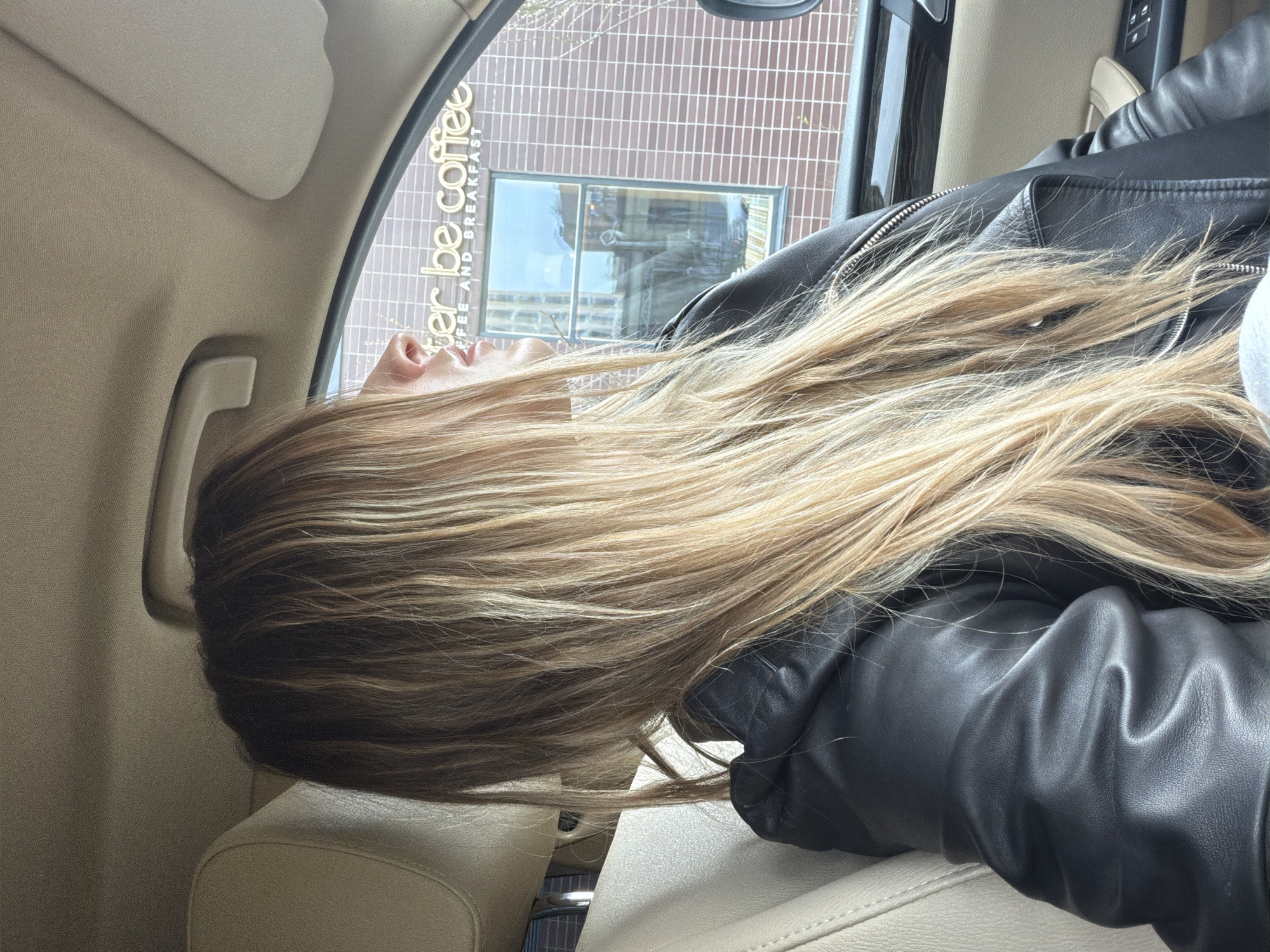 A woman is reclining in the back seat of a car. She has long, light brown and blonde hair, and she is wearing a black leather jacket. Outside the car window is a building with "er be coffee" printed on it. The full sign reads "Better be coffee/coffee and breakfast." Based on the sign, the image was taken in the city of Khimki, Russia. The car's interior is light beige.