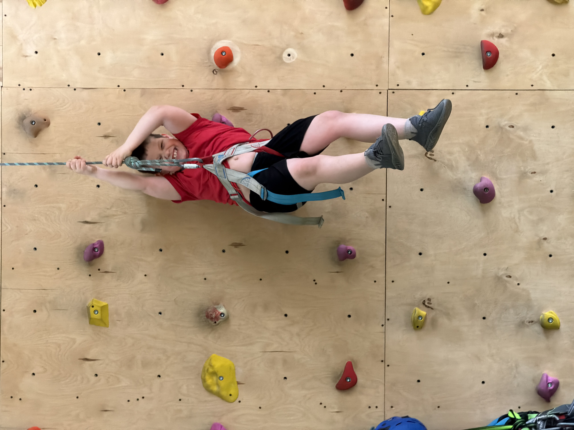 A child wearing a red t-shirt, black shorts, and grey sneakers is suspended horizontally from a climbing wall at a climbing gym. The child is secured with a climbing harness and rope. The climbing wall is made of light-colored wood and has various colored climbing holds attached to it. The photo was taken in Izhevsk, Russia.