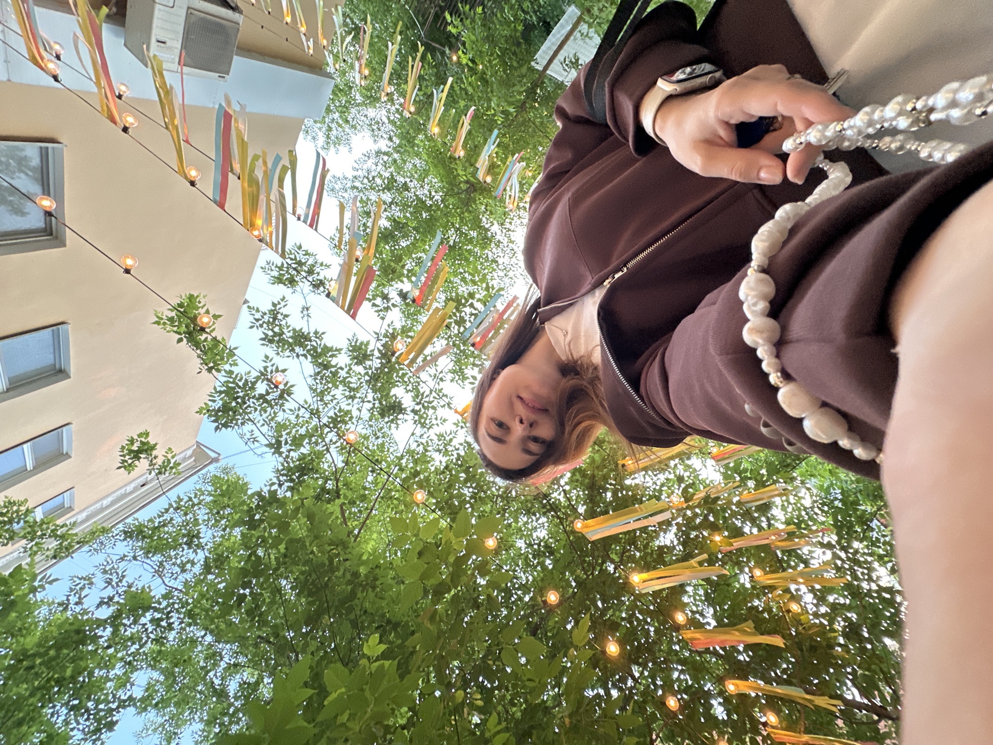 A low-angle, upward-facing image captures a young woman's selfie or close-up portrait in an outdoor setting in Perm, Russia. The woman's face, positioned towards the upper middle, looks directly at the camera with a neutral to slightly smiling expression. She wears a dark brown zip-up jacket over a lighter garment. Her right hand, adorned with a light-colored smartwatch, holds a decorative string of white, pearl-like beads.
The background is a vibrant mix of nature and urban elements, decorated with festive lighting. Lush green tree canopies fill much of the frame, with patches of bright sky visible through the leaves, indicating clear or partly cloudy weather. Numerous glowing string lights are strung across the scene, along with colorful, vertically hanging ribbons or streamers, suggesting a decorative or celebratory atmosphere, possibly for an outdoor event, park, or cafe area. To the left, a light-colored building with multiple windows and an external air conditioning unit is visible, grounding the scene in an urban environment. The overall impression is a pleasant, decorated outdoor space. No visible text is present.