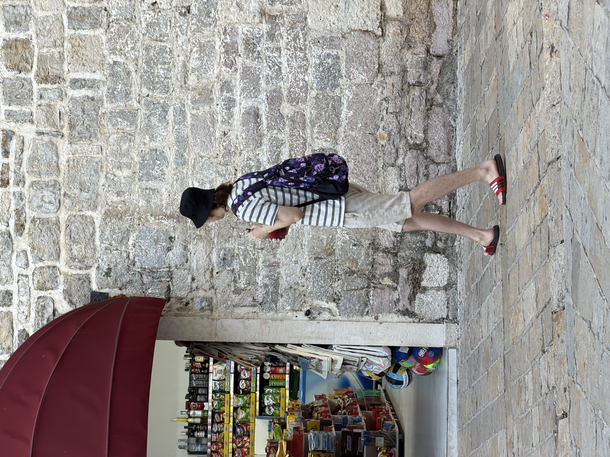 A person wearing a black bucket hat, a striped t-shirt, and light-colored shorts walks from left to right in front of a stone wall and a store. The person carries a backpack with a floral pattern and a red drink. The store, located in Budva, Montenegro, has a red awning and shelves stocked with various items, including bottles of alcohol and snacks. The ground is paved with cobblestones.
