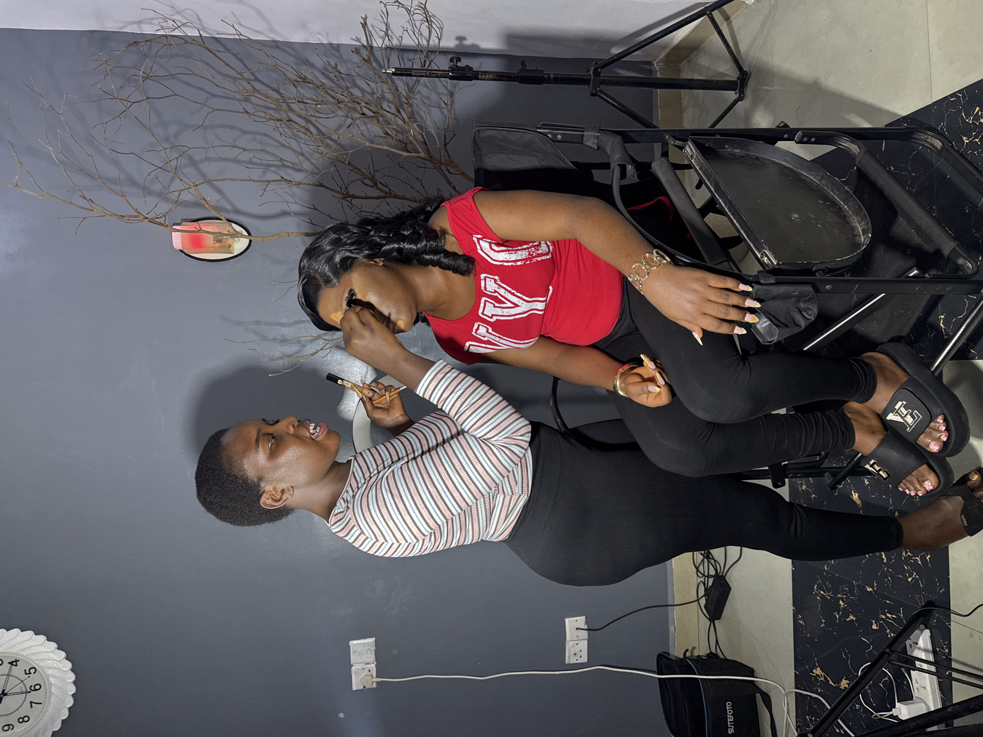 Two young Black women are in a room, likely a home or a studio, as evidenced by the makeup tools and equipment visible. One woman, wearing a red t-shirt with white lettering that appears to spell "V.I.K." or similar, is seated and appears to be having her makeup applied by the other woman. The woman applying makeup is wearing a striped long-sleeved shirt and black leggings. She is holding a makeup brush and is focused on her task.
The room has grey walls and a dark floor with a marbled pattern. A large branch, possibly a decorative element, is mounted on the wall behind the seated woman. There are also electrical outlets and cords visible, as well as a clock on the left side of the frame. The overall impression is that of a personal styling session or a behind-the-scenes look at makeup application. The lighting suggests it is daytime, or that the room is well-lit artificially. The women appear relaxed and engaged in the activity. There is no visible text other than on the woman's shirt and on some equipment in the background.