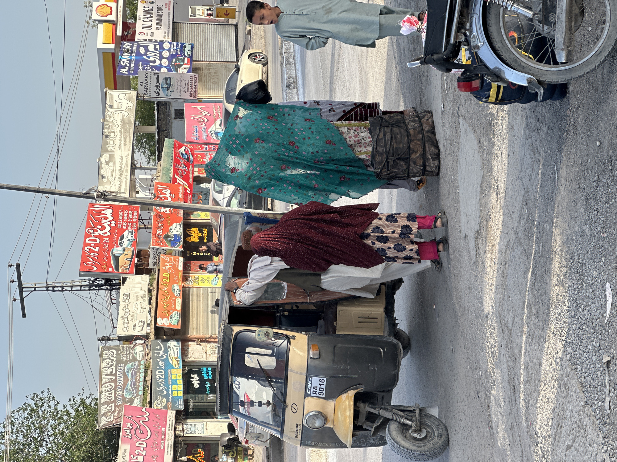 The image depicts a bustling street scene in Quetta City Tehsil, Pakistan, under bright, clear daylight. In the foreground, a beige and black three-wheeled auto-rickshaw (SAZGAR MNCAB, license plate RA 9016) is parked on the side of the road. A person wearing a maroon shawl over a patterned dress is leaning into the rickshaw's rear compartment, appearing to arrange or retrieve items. Another person, mostly obscured by a teal patterned dupatta, stands beside the rickshaw, while a third woman in a patterned dress is partially visible further left.
To the right of the rickshaw, a young man dressed in a light green shalwar kameez stands with an open-mouthed expression, looking towards the right side of the frame. A motorcycle's front wheel and handlebars are partially visible next to him. The street consists of a paved road with gravelly shoulders.
The background is densely packed with numerous commercial signboards, stacked vertically on utility poles and attached to storefronts. Many signs are in Urdu, with some English text, advertising various services. Recurring themes include "2D CAR SERVICE" and "Movers," often stating "24 گھنٹے آل پاکستان سروس گھرسے گھر تک" (24-hour all Pakistan service home to home). Specific business names visible include "KE MOVERS," "شاہ جی مووروز" (Shah Jee Movers), "المدينہ 2D," and "کورین ڈائی این 2 کار سروس." Phone numbers are prominently displayed on many signs. Other signs advertise "AL SARWARY SLEEPER & 2.0 CAR SERVICE," "DAEWOOEXPRESS," a "Shell" fuel station, and "FREE OIL CHANGE HAMMAD OIL STORE." The sky is clear blue, indicating sunny weather. The overall scene conveys a vibrant, commercial urban environment.