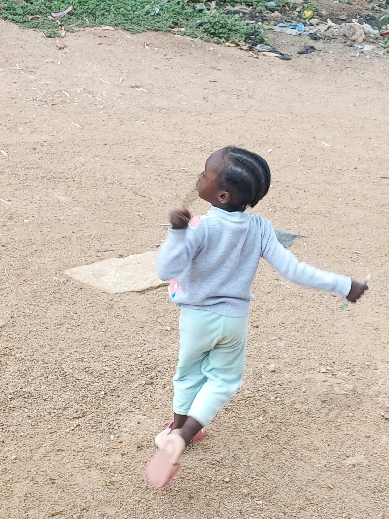A young girl is shown from the back. She has dark skin and her hair is neatly braided. She is wearing a light grey, long-sleeved shirt and light blue pants. She is wearing pink shoes. She is on a dirt road with trash and vegetation in the background. The photo was taken in Jos, Nigeria.