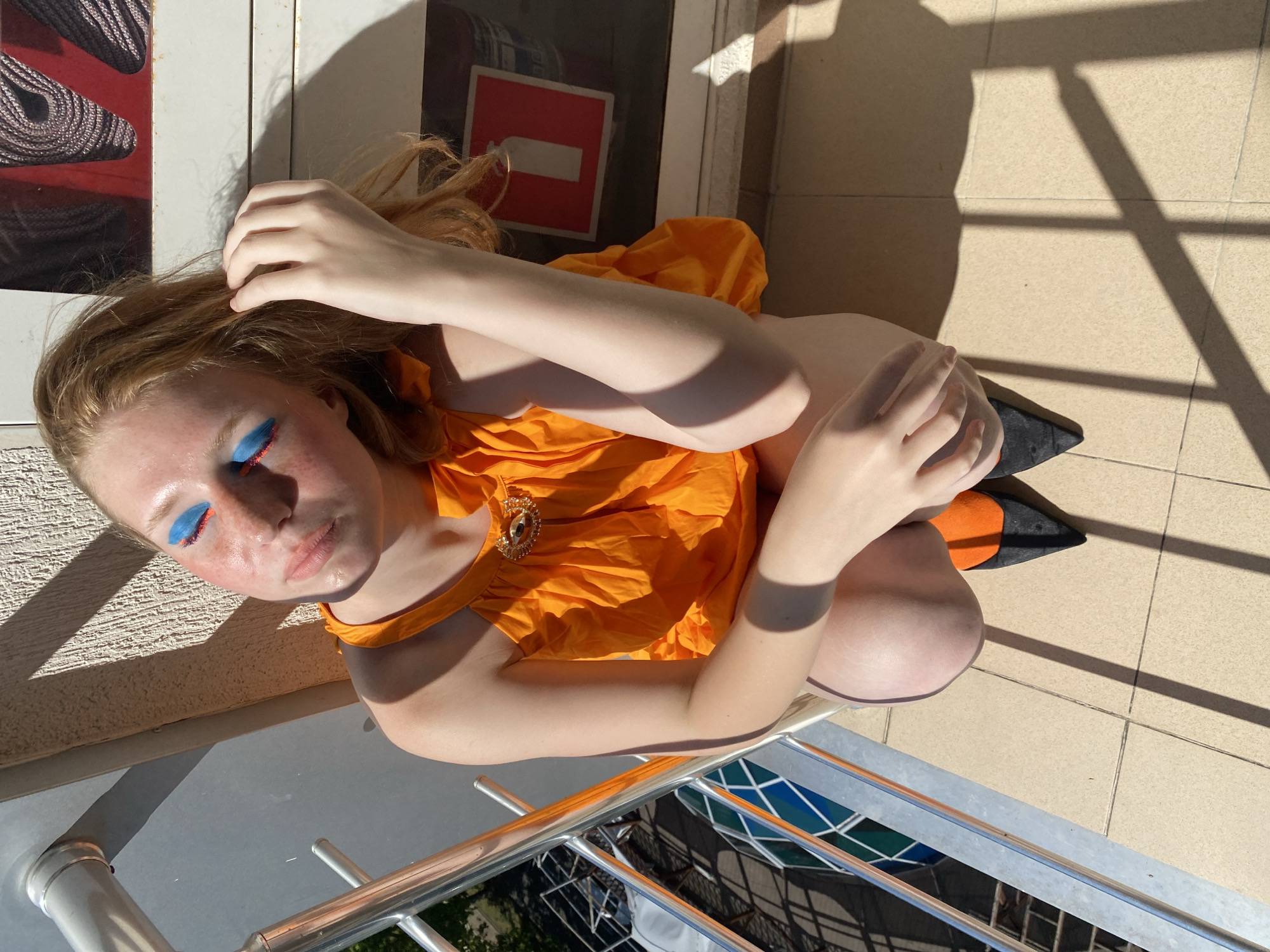 A young woman with long, reddish-blonde hair is photographed from a high angle outdoors. She is wearing a bright orange, ruffled top with a jeweled embellishment at the neckline, and dark pointed shoes with orange accents. Her eyelids are adorned with dramatic blue and orange eyeshadow. She is positioned on a tiled floor, with shadows cast from an unseen structure, suggesting it is a sunny day. The background includes a white vertical panel with a red and white square sign, and a glimpse of a metal railing with a reflective surface that shows a pattern of colored squares and some greenery. The overall scene suggests a playful and artistic pose taken during daylight hours. The sign in the background appears to be a no-entry or prohibition sign. The setting is likely a balcony or outdoor patio.