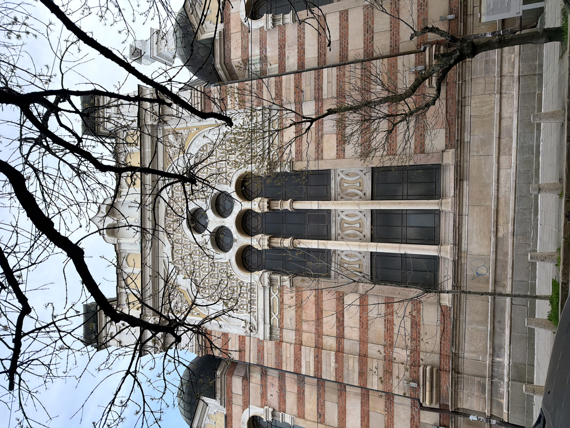 The image shows a building with striped brick and stone walls, featuring large arched windows and a dome. The building is partially obscured by bare tree branches in the foreground. The location appears to be urban, with elements of a sidewalk and street visible on the right.