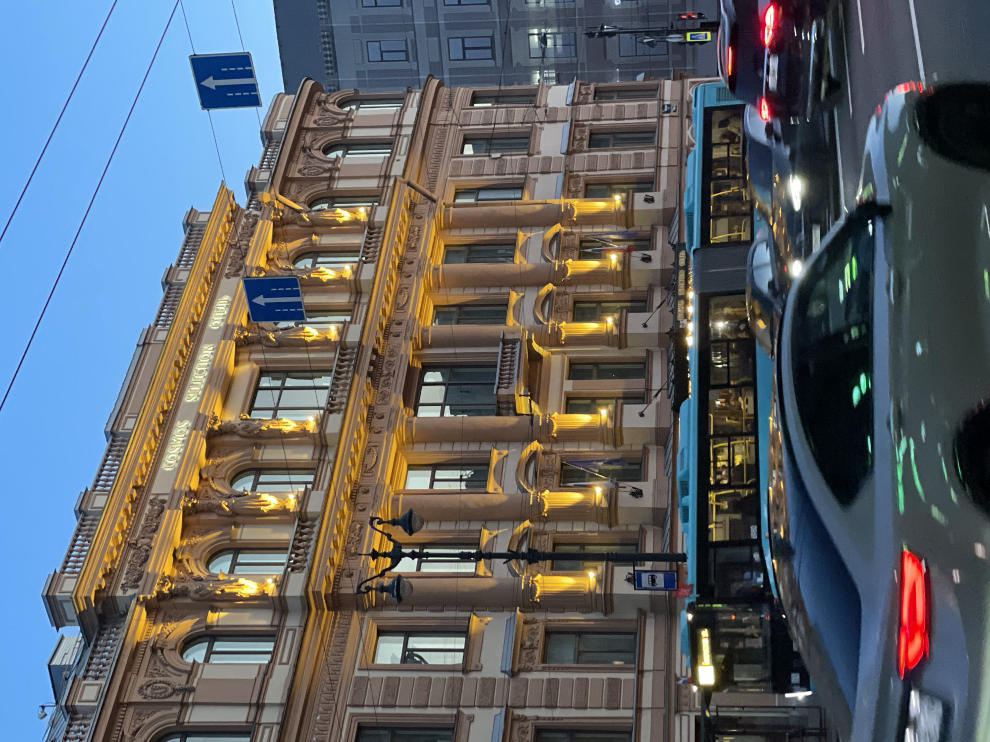 An evening urban scene in Saint Petersburg, Russia, showcases a grand, ornate building alongside a busy street. The multi-story building, likely a hotel or commercial establishment, features classical architectural elements such as decorative columns, balconies with railings, sculpted figures, and intricate moldings. Its facade is a light color, possibly beige or cream, and is warmly illuminated by golden lights, highlighting its details against the twilight sky.
Visible text on the building, illuminated in neon, reads "COSMOS HOTEL SOLUTION." Below this, the word "ОТЕЛЬ" (Hotel) in Russian is partially discernible.
The foreground and right side of the image depict a street with moving traffic. Several cars, including a white one in the immediate foreground and a dark one further down, have their lights on. A light blue city bus is also visible in the traffic flow. The road appears wet, reflecting the artificial lights from the vehicles and the building.
Two blue rectangular traffic signs with white left-pointing arrows are prominent. One is suspended high above the street by wires, and another is attached to the building at a lower level, indicating traffic direction or restrictions. A standard blue bus stop sign is also visible near the bus. The sky above is a clear, deep blue, signifying twilight or early evening, creating a contrast with the brightly lit building and vehicle lights. The architecture and traffic signs are consistent with the provided location context of Saint Petersburg.