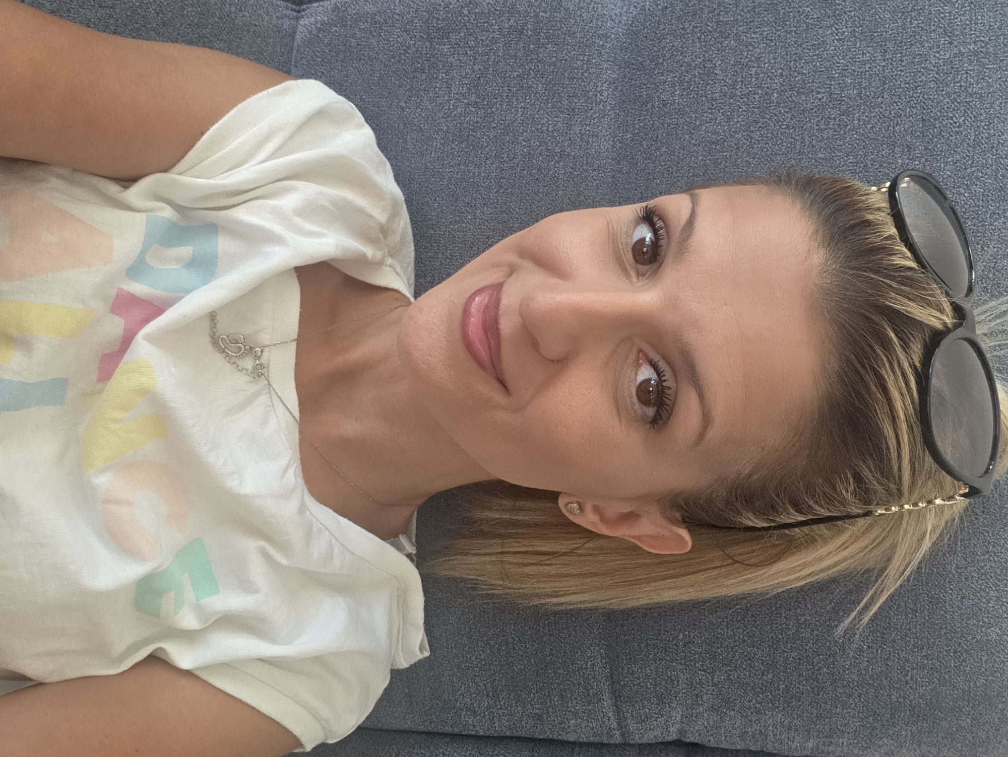 A woman with blonde hair pulled back into a ponytail and sunglasses perched on her head is looking at the camera and smiling. She is wearing a white t-shirt with colorful lettering on it and a silver necklace. The woman is sitting on a grey couch. The image was taken in Munich, Germany.