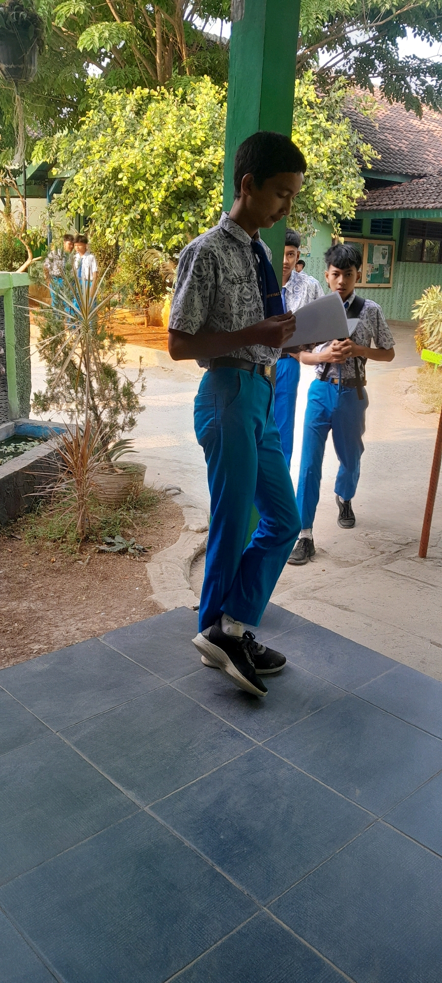 In Tahunan, Indonesia, a young male student, wearing a white patterned shirt, blue tie, and bright blue trousers, stands on a tiled porch, holding a document. His dark hair is short, and he is looking down at the paper. Behind him, two other male students in similar uniforms are also visible. One, with dark hair, wears a white patterned shirt, blue tie, and blue trousers, and is looking towards the camera. Another student, further back, is mostly obscured. The environment appears to be outdoors, with lush green trees and foliage in the background, suggesting a school or public area. A building with a brown tiled roof is visible behind the students, and a green pillar stands to the right of the central student. The foreground shows a dark blue tiled walkway. The scene is lit by natural daylight, casting soft shadows.