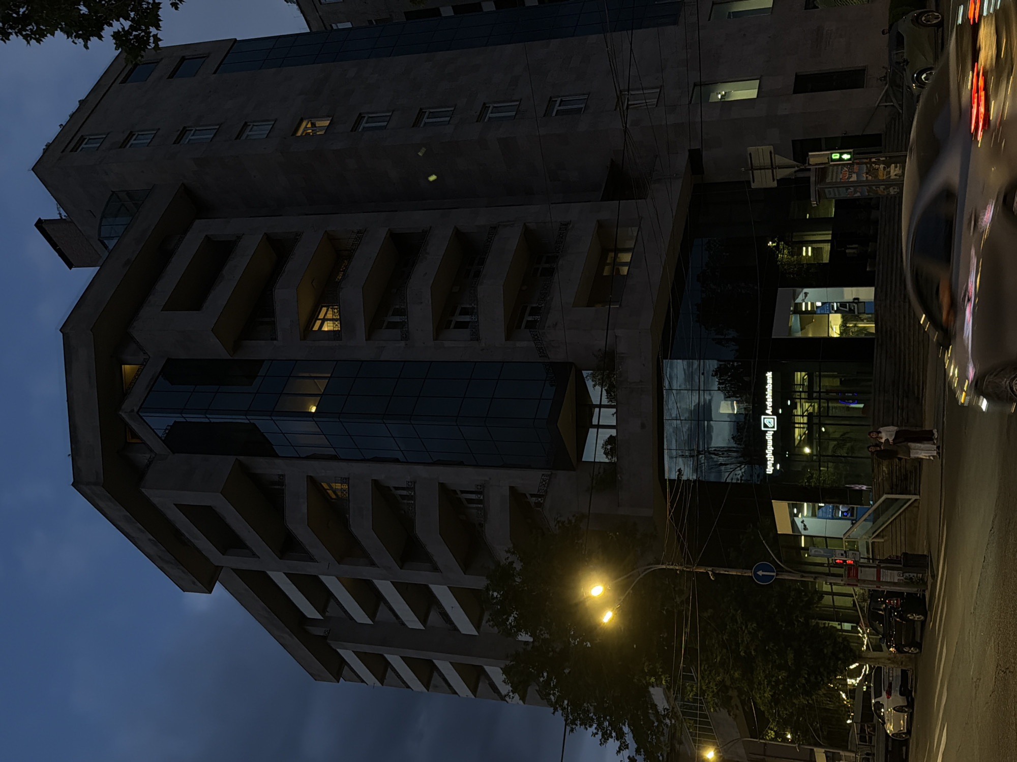 This image captures an urban street scene during the evening or night. The primary subject is a multi-story, modern building with a distinctive architectural style, featuring a concrete facade with angular, recessed windows or balconies, and sections of dark glass. Several windows within the building emit warm yellow light, indicating interior activity.
On the ground floor, the building features large glass panels reflecting the street and artificial lights. Visible on the glass facade is text in both Latin and Armenian scripts, reading "Ardshinbank" (Արդշինբանկ).
In the foreground and to the right, a street runs parallel to the building. Parked cars are visible along the curb, and reflections of vehicle lights, notably red and white streaks, suggest traffic or passing cars. Two people are standing on the sidewalk near the building's entrance. Streetlights illuminate the scene with a warm glow, casting light on parts of the building and the street. Trees with dark foliage are scattered along the street and partially obscure parts of the building and sky. A blue road sign with a white arrow pointing left is visible on a pole near the sidewalk, along with what appears to be a bus stop or public transport information display. The sky is a dark blue-grey, characteristic of dusk or nighttime.