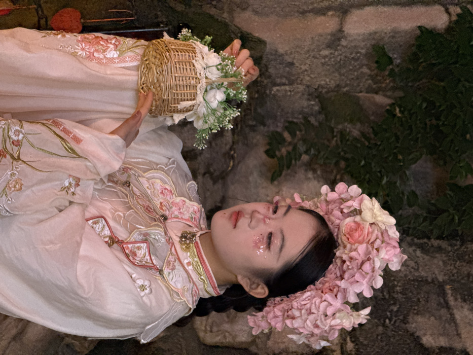 A young individual, presumed female, of East Asian ethnicity, is depicted in a waist-up, profile portrait against a textured rock wall partially obscured by dark green foliage. The scene is located in Yuzhong District, China. The individual wears an elaborate headpiece constructed from numerous pink and some cream-colored artificial flowers, including what appear to be hydrangeas and roses. Her dark hair is pulled back, partially visible beneath the headpiece. Her facial makeup includes glitter applied to the upper cheeks and a pinkish hue around the eyes. She is dressed in a light-colored, possibly pale pink or white, traditional Chinese-style garment featuring a high collar with pink and gold trim and a central golden ornament. The garment's bodice and wide sleeves are adorned with intricate floral embroidery in shades of pink, green, and gold. A decorative pink and white pendant-like element hangs from the front of the garment. She holds a small, light brown woven wicker basket with both hands, positioned slightly forward. The basket contains a cluster of artificial white and green flowers and foliage, and its rim is trimmed with cream-colored lace. A red, heart-shaped object is partially visible hanging on the left side of the frame.
