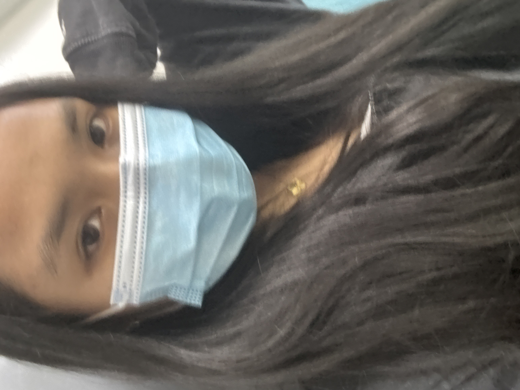 The image shows a close-up of a young woman wearing a light blue surgical mask and a dark gray hoodie. Her dark, long hair frames her face and cascades over her shoulders. She is wearing a small, gold pendant necklace. The background is out of focus, but appears to be an indoor setting with a light-colored wall and a hint of a blue object on the right. The lighting is soft and diffused. No specific location within Davao City can be identified from the image.
