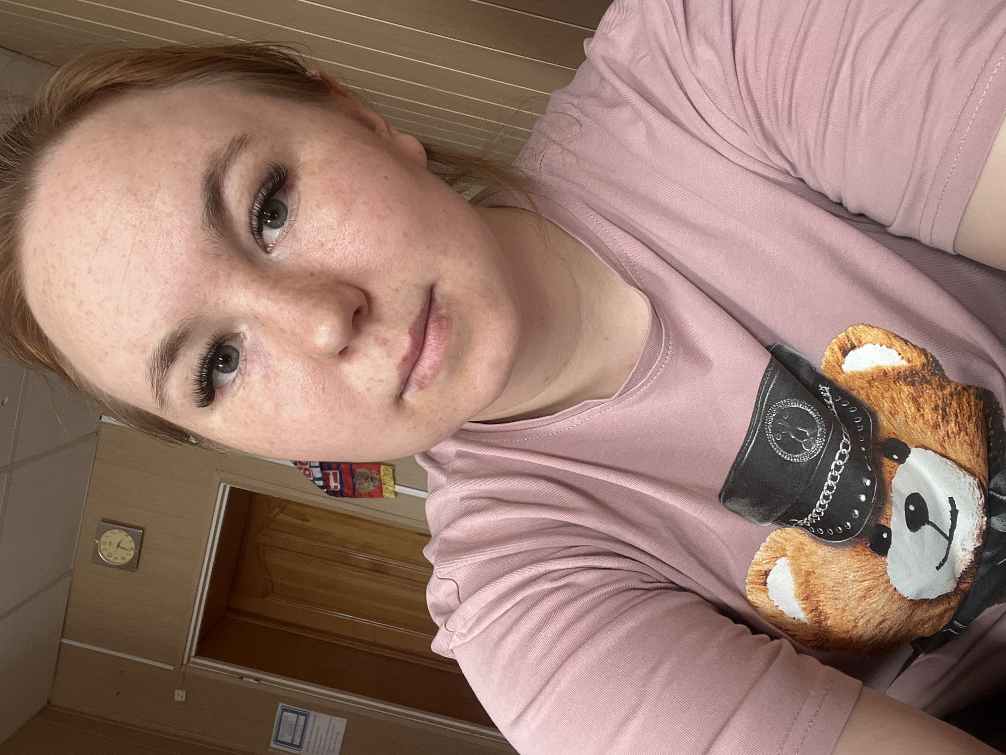 A woman with fair skin, freckles, and auburn hair, is pictured in a selfie. She has long eyelashes. She is wearing a pink T-shirt with a teddy bear print wearing a black cap with silver chains. A wall with a clock is behind her. A wooden door is visible. The clock shows it is after 9. Based on the file name, the location is Chaikovsky, Russia.