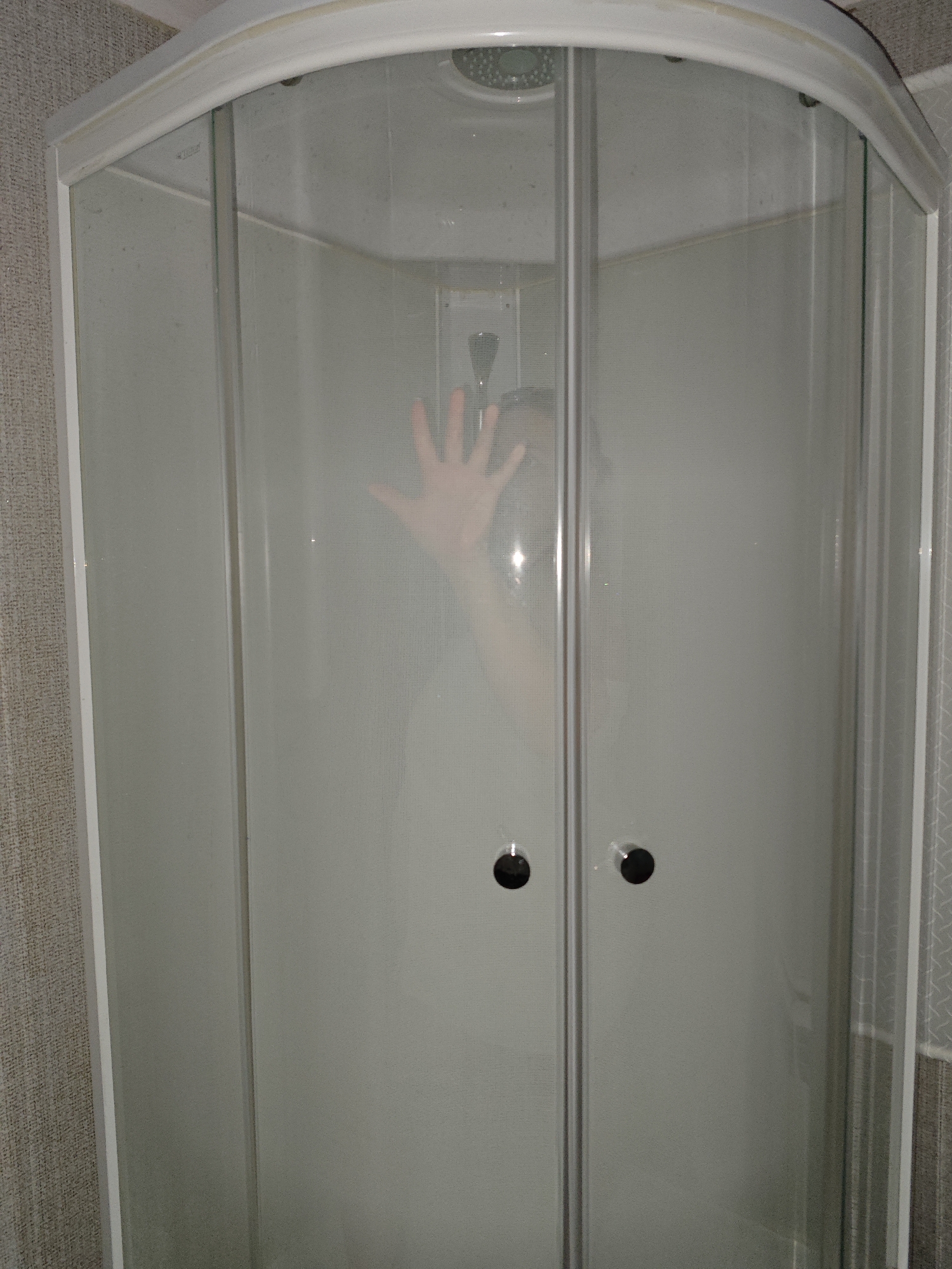 A steamy shower stall is visible, with an indistinct figure pressing a hand against the translucent glass. The shower has a curved design with white framing. The surrounding wall has a textured, neutral-colored wallpaper. The location, Фоки, Russia, cannot be verified from the visual information provided.