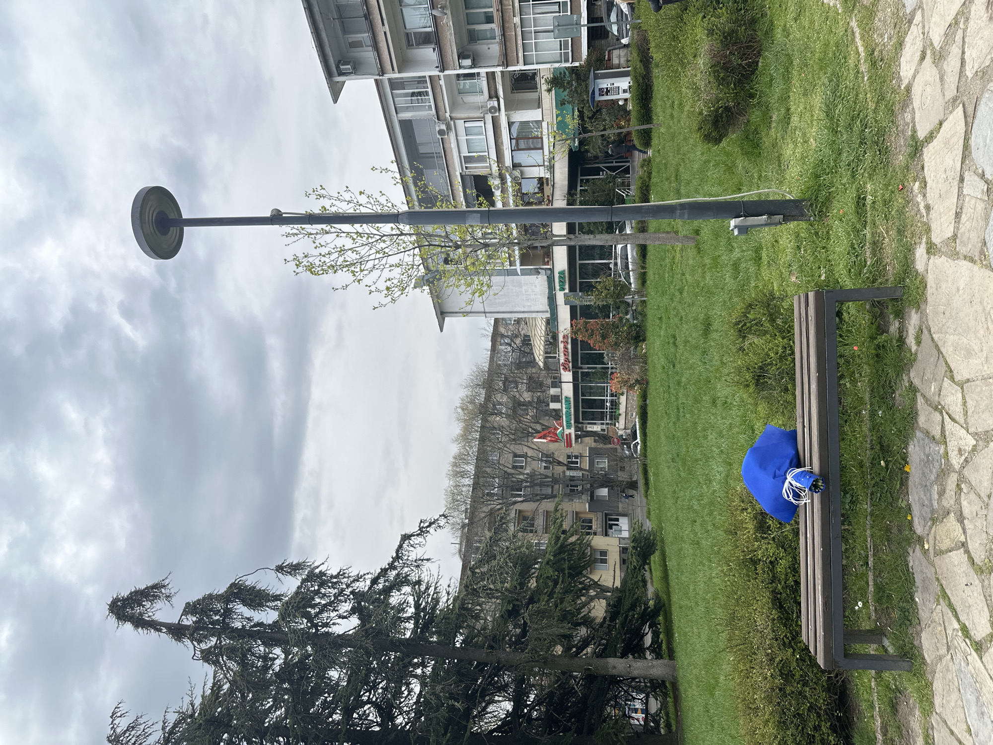 This image captures a quiet urban park scene under an overcast sky during the daytime. The foreground features a stone path on the right, bordering a vibrant green grassy area with scattered trees. A dark, tall lamp post stands prominently towards the left. On the right, a wooden park bench holds a bright blue bag, possibly a backpack, tied with white strings.
In the mid-ground, a variety of trees are visible, including mature coniferous trees with dense foliage and deciduous trees beginning to sprout fresh, light green leaves, indicating spring. Beyond the green space, a line of multi-story apartment and commercial buildings forms the background. These buildings exhibit various architectural styles, featuring numerous windows, balconies, and air conditioning units.
Visible text includes "PIZZA" on one building facade. Another building in the distance displays a red star logo, possibly accompanied by text that appears to be "БУРГЕР" in Cyrillic. No people or specific activities are depicted, giving the scene a calm, everyday ambiance. The cloudy sky suggests a mild or cool day. The overall setting is typical of a European city park integrated within a residential and commercial area.