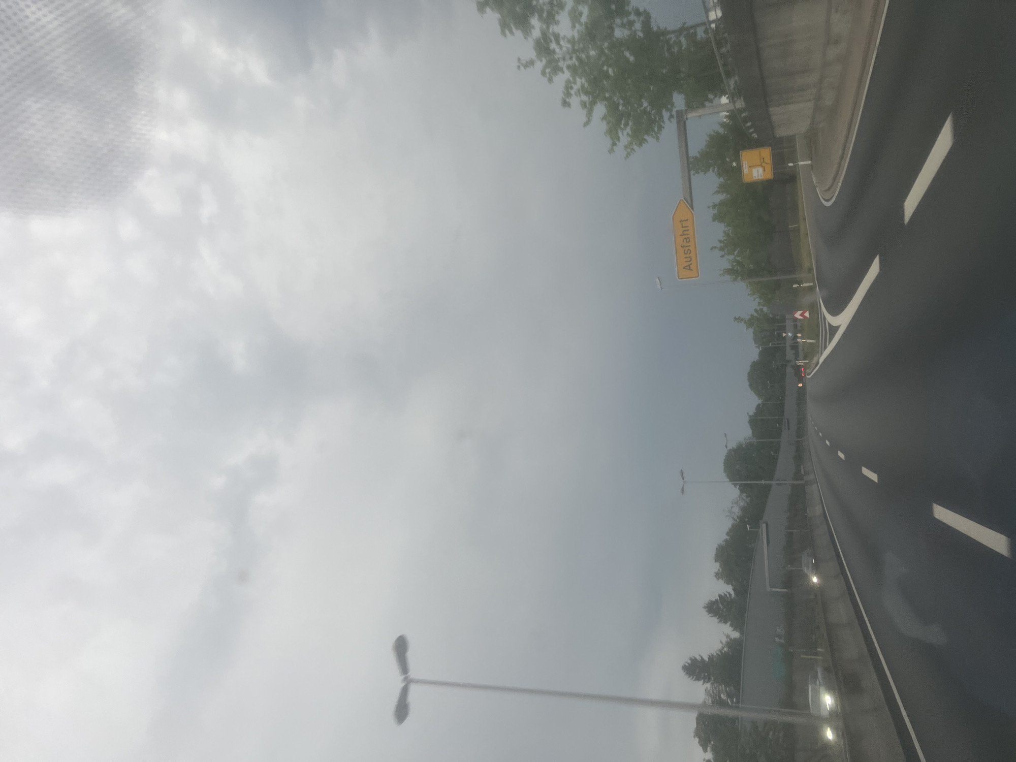 The image shows a view from a car on a highway in Augsburg, Germany, on a cloudy day. The sky is overcast, and the overall lighting suggests it might be late afternoon or early evening. A yellow road sign with black text indicating "Ausfahrt" (German for "exit") is visible on the right side of the road. The highway has multiple lanes and is wet, possibly due to rain. Trees line the side of the road, and a concrete barrier runs along the right edge. Several streetlights are also visible. The image appears to have been taken from the driver's or passenger's seat, looking forward and slightly to the right.