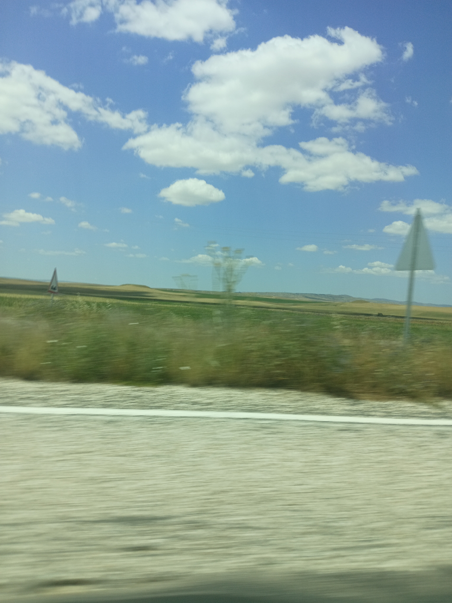 The image is a blurred, horizontal shot taken from a vehicle, likely on a sunny day. The sky is a clear blue with scattered white clouds. In the distance, rolling hills are visible, covered with fields of green and brown vegetation. There are a few signs visible, including a triangular warning sign on the left and a white triangular sign on a pole on the right. The foreground shows a blurred road surface, with a white line running horizontally across it. The image does not show any people or specific city landmarks in Balâ, Turkey.