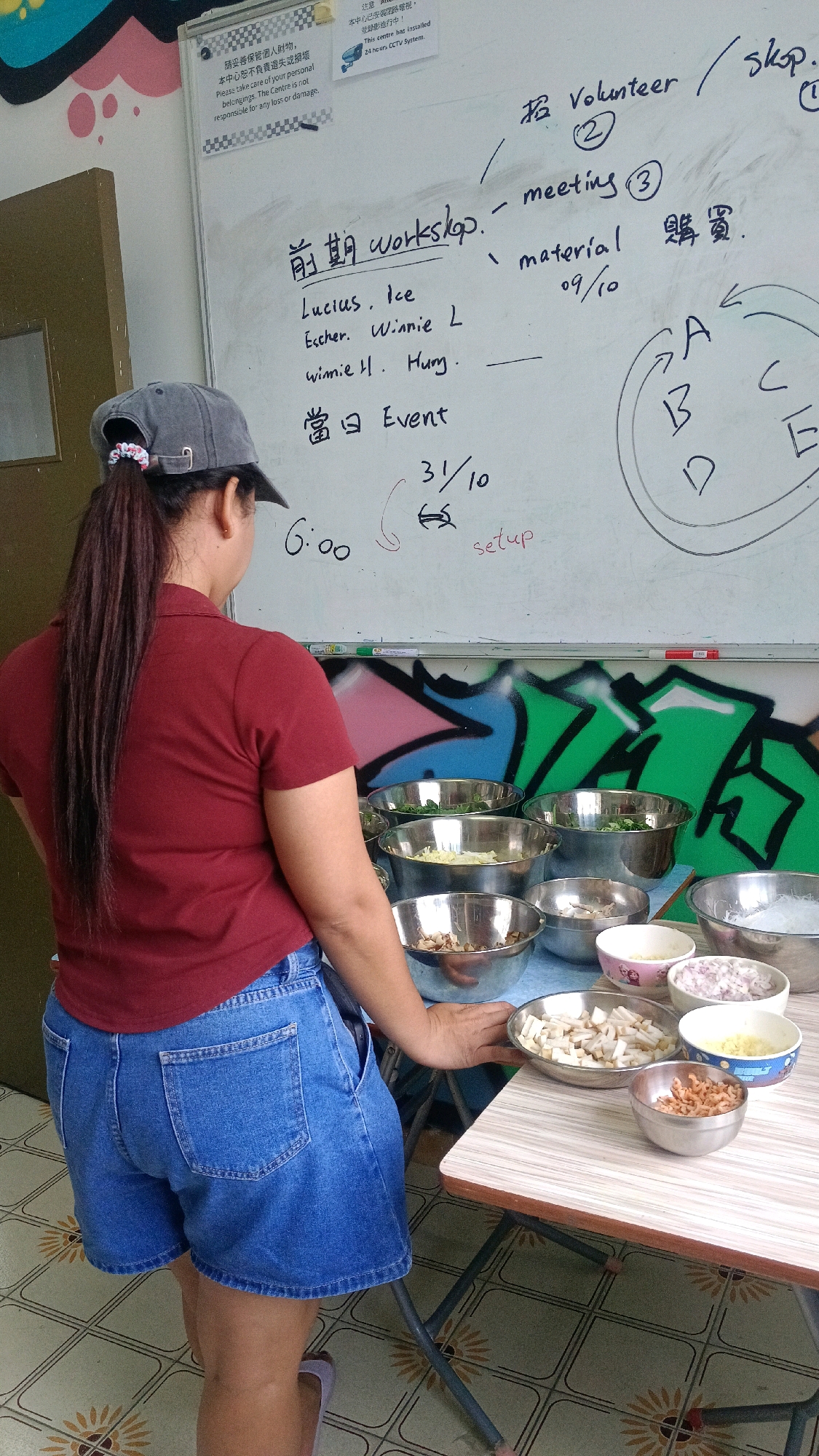 The image shows a woman wearing a red polo shirt and denim shorts standing in front of a whiteboard covered in handwriting. The whiteboard has titles like "Workshop," "Volunteer," and "meeting." Below the workshop title, names and dates are listed, including "Lucius Ice," "Escher Winnie L," "winnie H. Hung," and "09/10." A section labeled "Event" is also visible. To the right of the whiteboard, a circular diagram with letters A, B, C, and E is drawn. In the foreground, a table is set with various bowls of food ingredients, including chopped vegetables, nuts, and noodles. The room appears to be an indoor space with tiled flooring and graffiti on the wall behind the whiteboard. The overall impression is that of a preparation or planning session for an event or workshop. The presence of English text and the style of the whiteboard suggest a potential location in Hong Kong, China, as it's a common practice for bilingual signage and notes in such settings.
