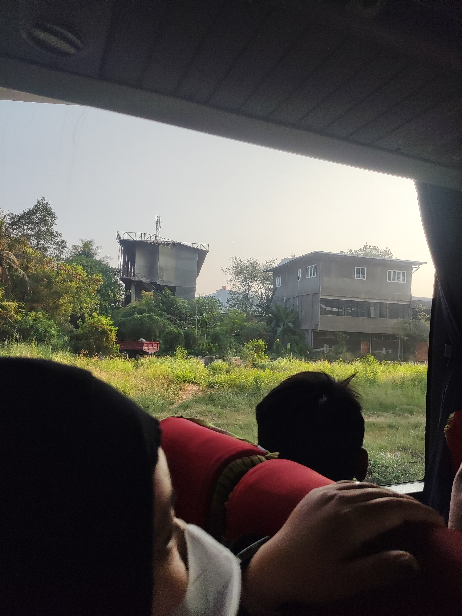 The image, taken from inside a vehicle, looks out onto a scene in Sukaresmi, Indonesia. In the foreground, the backs of several people are visible, suggesting they are passengers. One person wears a red vest. Outside the vehicle, a grassy field stretches towards buildings and lush vegetation. To the left, a partially constructed building with a metal frame and a tower stands amidst trees. To the right, a two-story building with multiple windows is visible. A red truck is parked in the grassy area between the buildings. The sky is overcast, and the overall lighting suggests it is daytime.