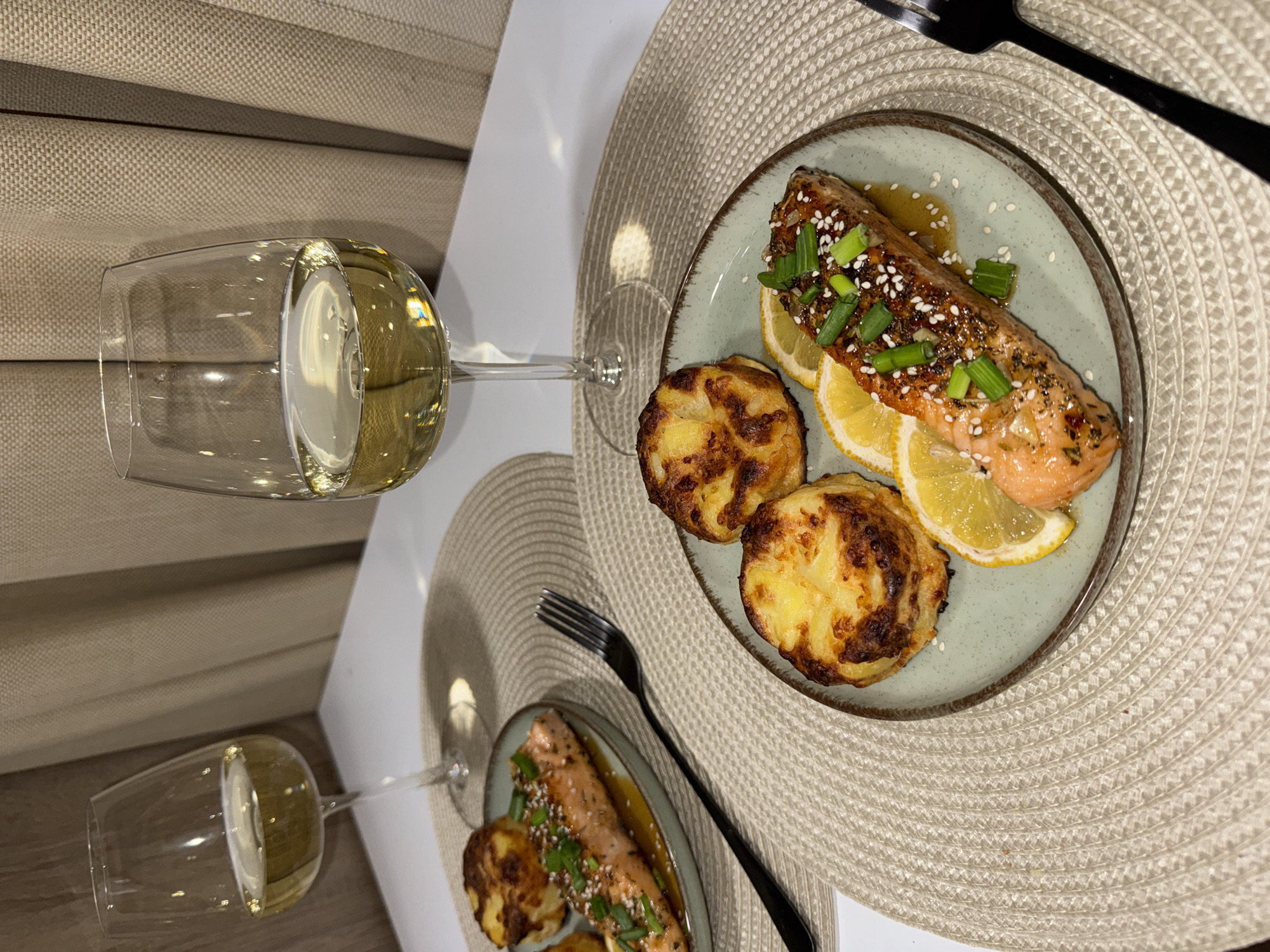 The scene depicts a meticulously arranged dinner for two in an indoor setting in Sofia, Bulgaria. Two individual place settings are positioned on a white table. Each setting includes a round, light beige woven placemat. The foreground setting on the right features a round, light green plate with a brown rim. On this plate, a portion of baked salmon, garnished with chopped green onions and white sesame seeds, rests on three thin lemon slices and a light sauce. Two individual, round, golden-brown baked potato gratin dishes accompany the salmon. A black metal fork is placed to the right of the plate. Behind this setting, a clear stemmed wine glass holds a portion of white wine. A second, partially visible setting on the left mirrors the first, displaying a salmon dish, at least one potato gratin, a black fork, and a wine glass containing white wine. The background consists of a plain light-colored wall and textured beige curtains on the right side. The overall lighting is warm, artificial indoor light.