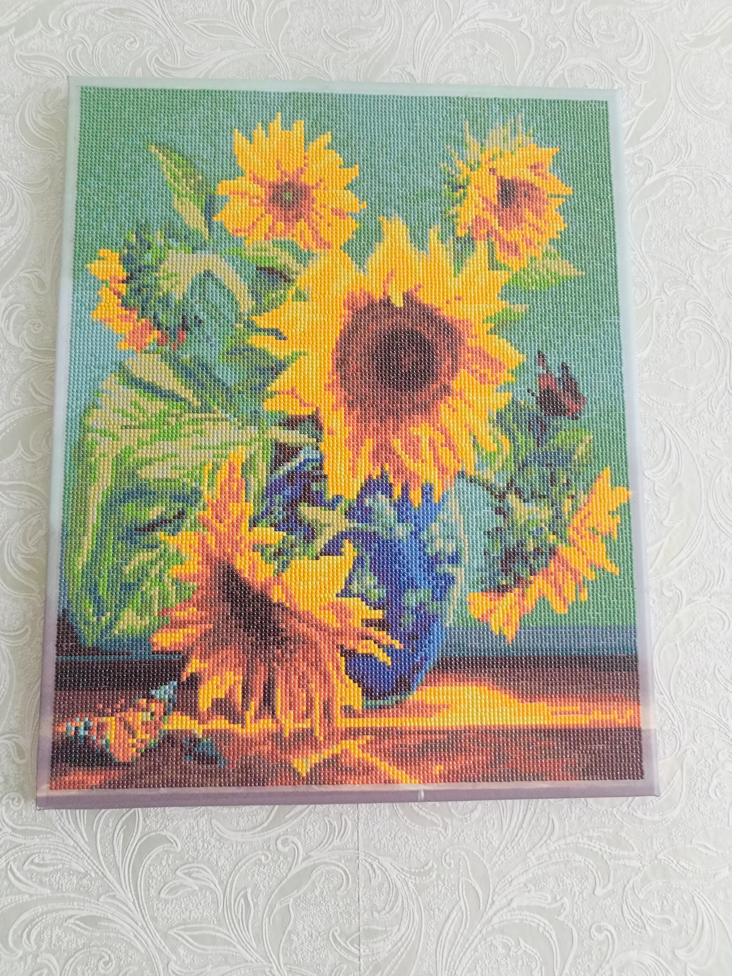 The image shows a diamond painting of sunflowers in a blue vase. The painting is framed and appears to be hanging on a textured white wall. The sunflowers are depicted in various stages of bloom, with bright yellow petals and dark brown centers. Green leaves and stems surround the flowers, and a small brown butterfly is perched on one of the leaves. The background of the painting is a gradient of green, transitioning to a warm orange and brown at the bottom, suggesting a sunset or warm lighting. The diamond painting technique creates a textured, pixelated effect. The location of this image within the city of Vitsebsk, Belarus, cannot be determined from the provided visual information.