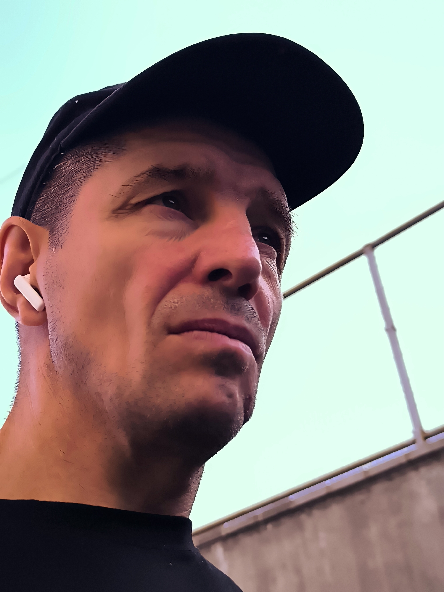 A close-up, low-angle shot shows a man wearing a black baseball cap and a black t-shirt. He has short, dark hair and is looking upwards and to the right. A white wireless earbud is visible in his left ear. The background consists of a light blue sky and a blurred structure with horizontal and vertical bars. The image is digitally enhanced, giving it a vibrant, almost painted quality. The location is specified as the city administration of Qarağandı, Kazakhstan.