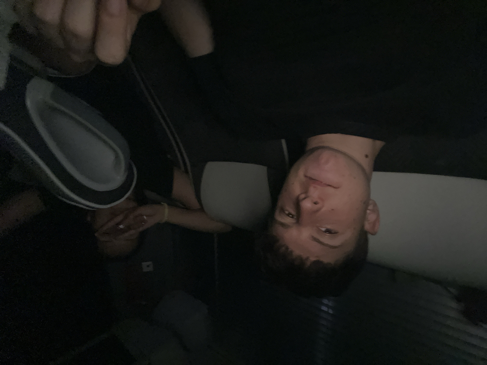 Two people are in a dimly lit indoor space. One person is facing the camera. They are wearing a dark-colored shirt. Another person is near the bottom of the image; their face is partially obscured by their hand. There is a circular object with a lighter colored rim near the top left of the image. The location is not recognizable as Dinkata, Bulgaria.