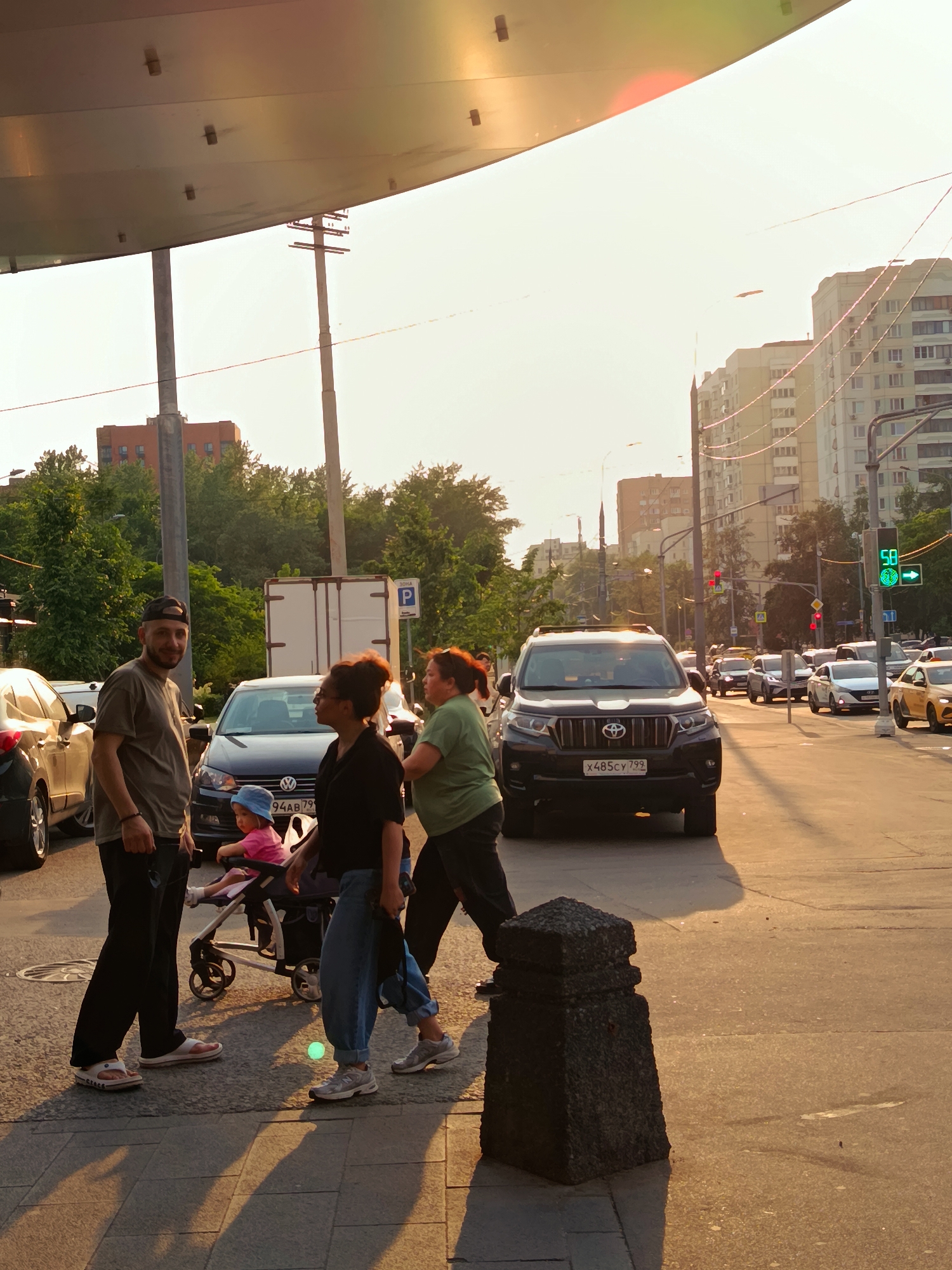 In Moscow, Russia, at sunset, a man in a grey t-shirt and baseball cap, and a woman in a green t-shirt and black pants, stand on a sidewalk near a covered walkway. A toddler in a pink outfit and blue hat sits in a stroller between them. To their right, another woman in a black shirt and jeans walks past. A dark SUV, a black Volkswagen sedan, and other vehicles are on the street in front of them, with traffic lights indicating a green signal. Buildings and trees line the street.