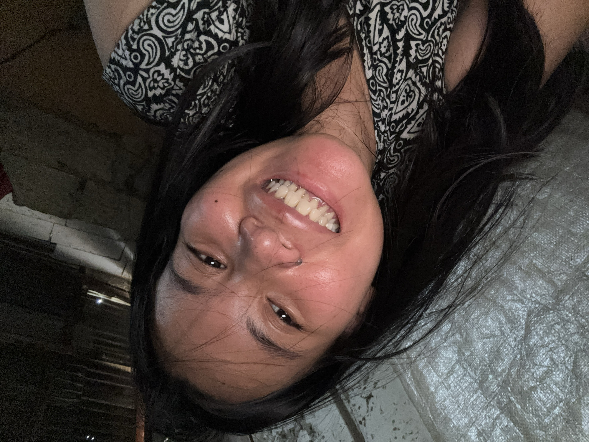 A young woman with long, dark hair and a black and white patterned top smiles at the camera in a dimly lit indoor setting. Her face is close to the camera, and her eyes are open. Her teeth are visible in her smile, and she has a nose ring. The background appears to be a wall made of rough, textured material, possibly concrete or stone, with some wooden beams visible at the top. To the left, a large, crinkled, silver-colored sheet or tarp is present, suggesting a rustic or unfinished environment. The lighting is low, with some highlights on her face and hair, and the overall scene is underexposed, creating shadows and obscuring finer details of the surroundings. The image was taken in East Jakarta, Indonesia.