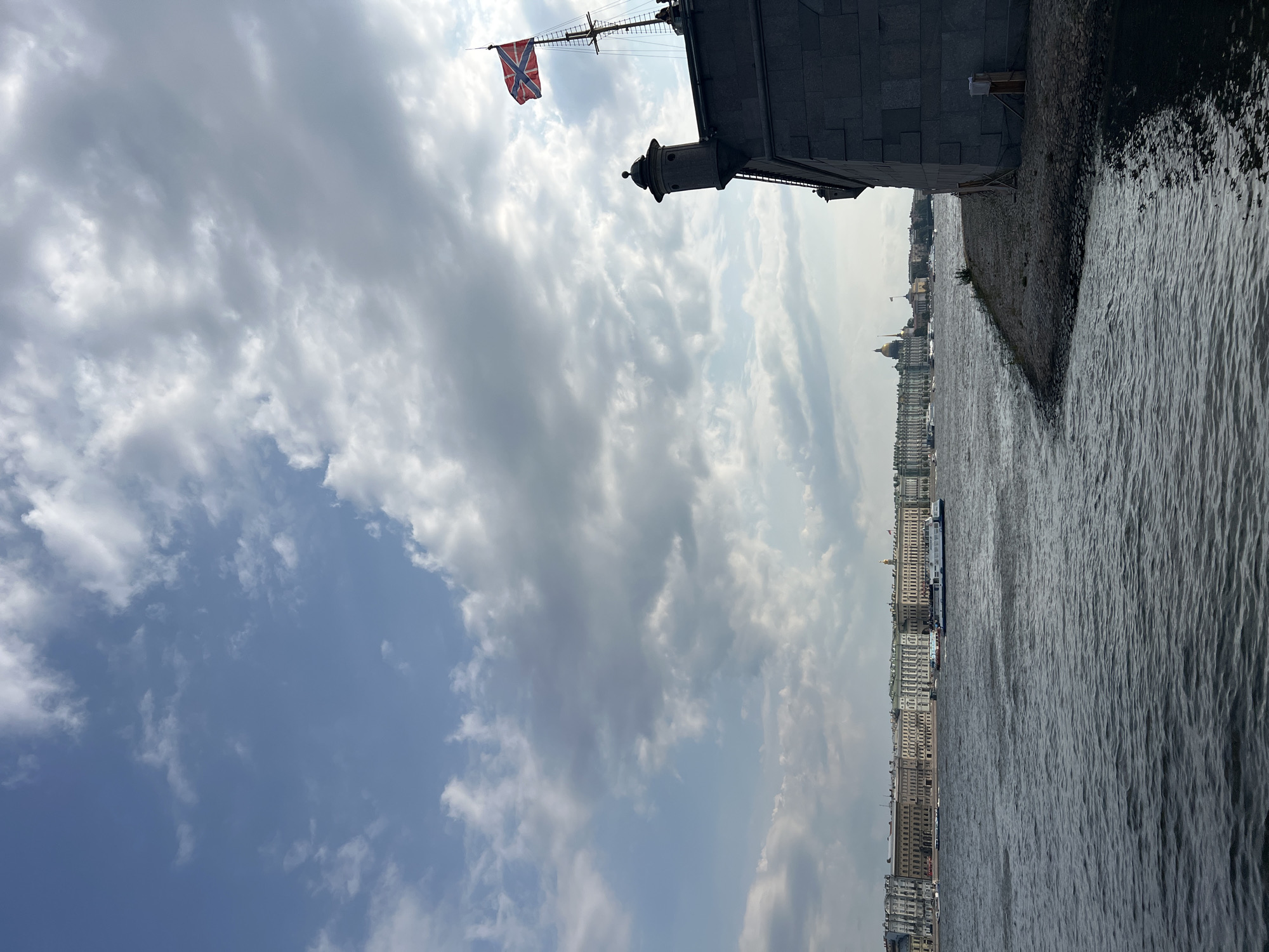 The image shows a view of the Neva River in Saint Petersburg, Russia, on a cloudy day. In the foreground, a section of a stone embankment and a body of dark water are visible. On the right side of the image, a large stone structure, likely part of a fort or citadel, juts out towards the river, topped by a flagpole with a flag. In the background, across the river, a long, low-rise cityscape with historic buildings stretches along the waterfront. Several boats, including a large white passenger vessel, navigate the river. The sky is filled with a mix of white and gray clouds, with patches of blue sky visible. The overall scene conveys a sense of historical architecture and riverine urban landscape.