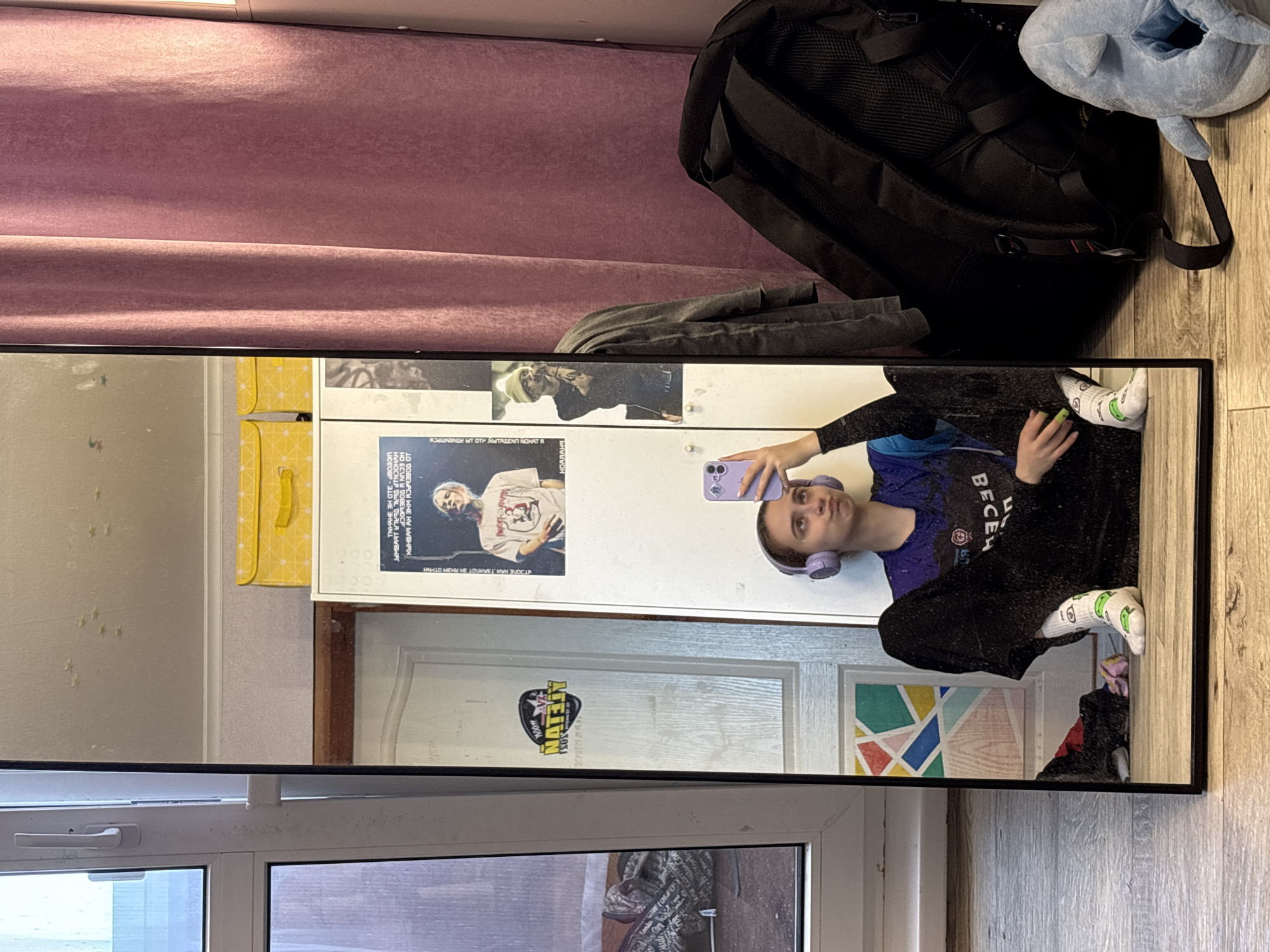 The image shows a mirror in a room. In the mirror's reflection is a person taking a selfie with a purple phone while wearing purple headphones, a blue and black shirt with the word "BECEM" on it, and black pants. They are wearing white socks with a green avocado print.
The background reflected in the mirror includes posters on a white wall, a yellow wallet with a pattern, a white door with a "NAFTAN" sticker, and a colorful geometric artwork. A window with closed blinds is at the bottom of the reflection.
Outside the mirror's reflection, a purple curtain is at the top of the frame, along with a black backpack and a blue stuffed animal. The floor is light brown wood.
Based on the OCR, a poster is in Russian and may have the words "Прикамский, Russia" on it.
