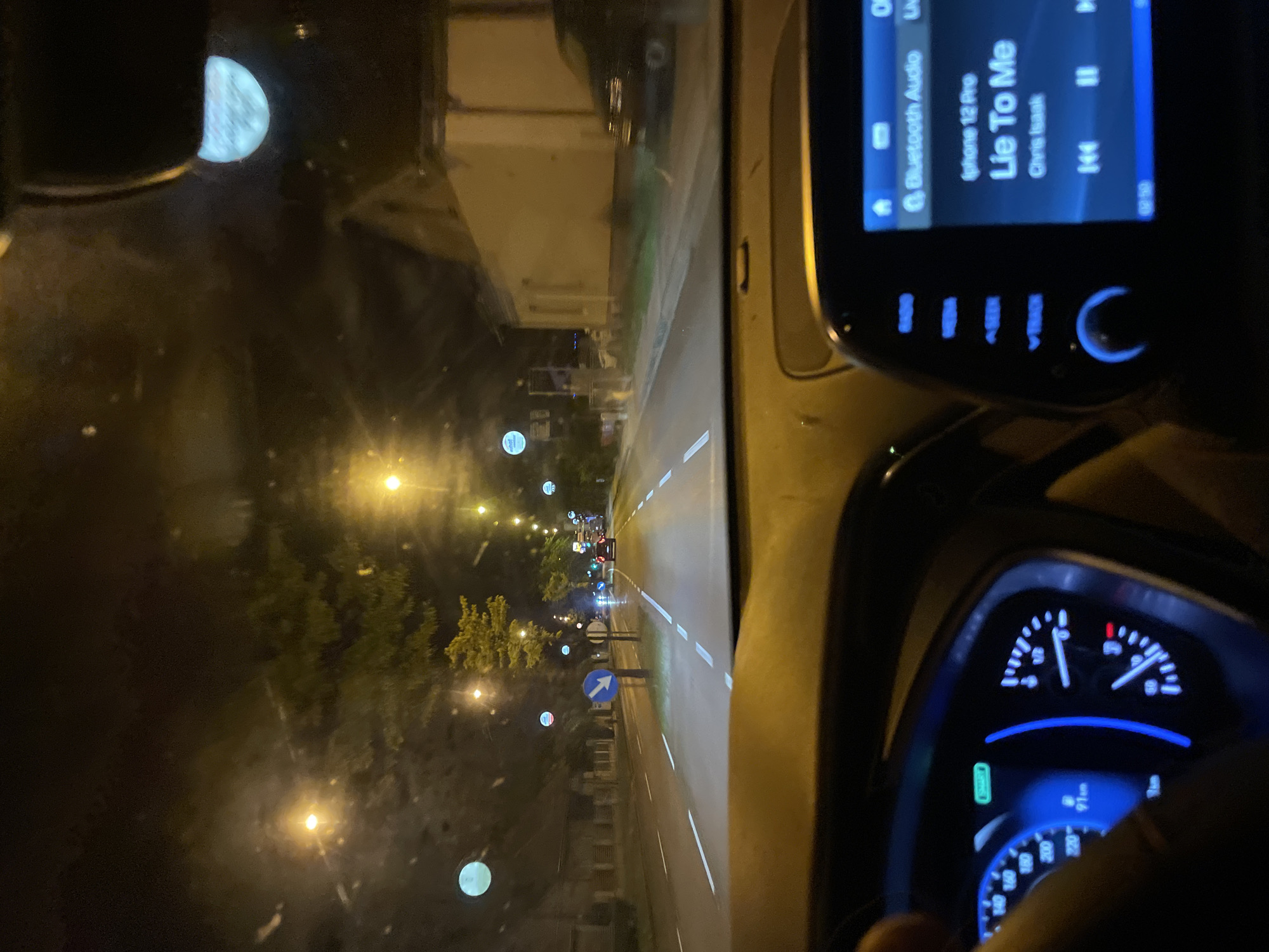 The image shows the view from inside a vehicle driving on a city street at night. The vehicle's dashboard is visible, with the speedometer illuminated and displaying approximately 50 km/h. A screen indicates the song "Lie To Me" by Chris Isaak is playing from an iPhone 12 Pro connected via Bluetooth. Rain is visible on the windshield. The street outside is illuminated by streetlights, and there are buildings and trees lining the road. There are other cars ahead on the road. A street sign with a blue arrow pointing to the right is visible. The location is indicated as Unknown, Unknown.