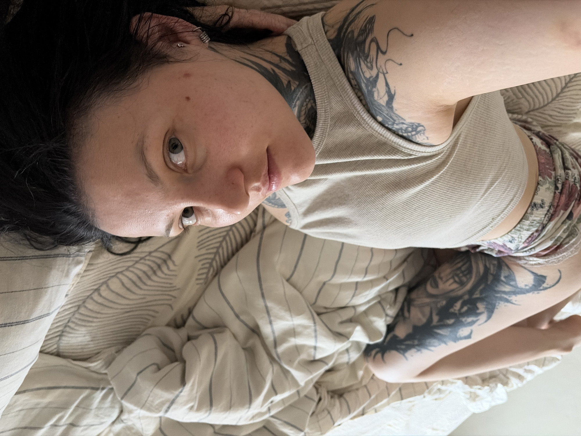 A fair-skinned woman with dark hair is lying on a bed. She has tattoos on her upper chest, neck, and right arm. She's wearing a beige ribbed tank top and floral patterned shorts. The bed has beige and gray striped sheets and a white ruffled edge. The woman's face is visible with light eyes, and multiple piercings are visible in her left ear. The location is not discernable from the provided image.