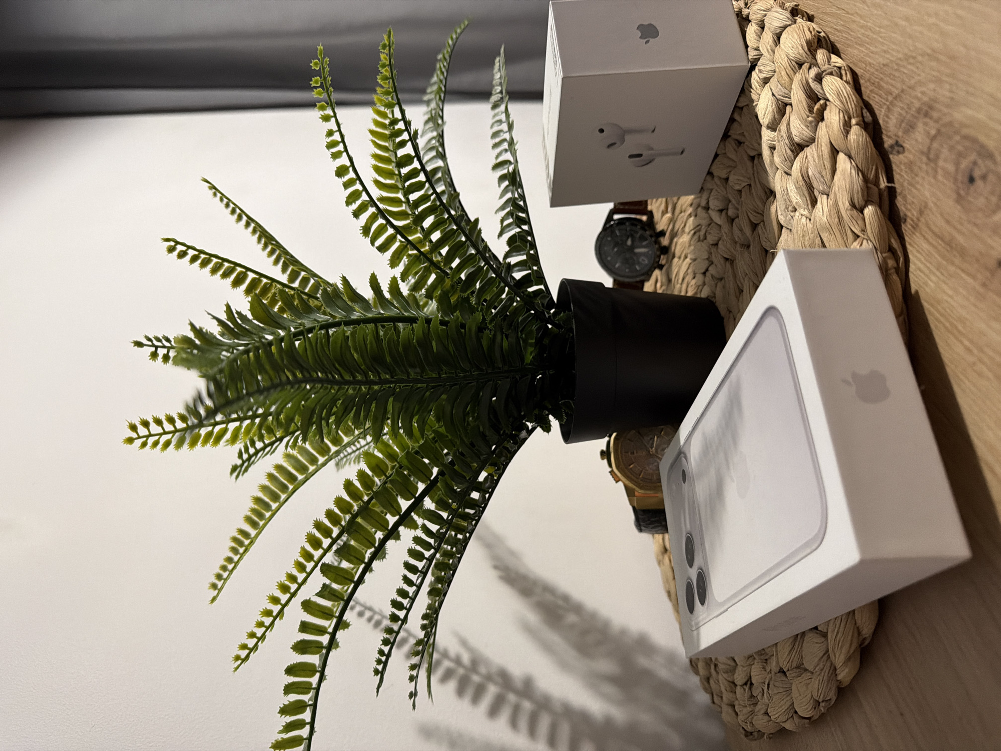 An artificial fern plant in a black pot sits on a woven placemat. To the left of the plant is a white box containing an iPhone, and to the right is a white box with an image of AirPods on it. Two watches are visible behind the plant. The items are arranged on a wooden table against a white wall. The image does not contain information about the city of Sosnowiec, Poland.