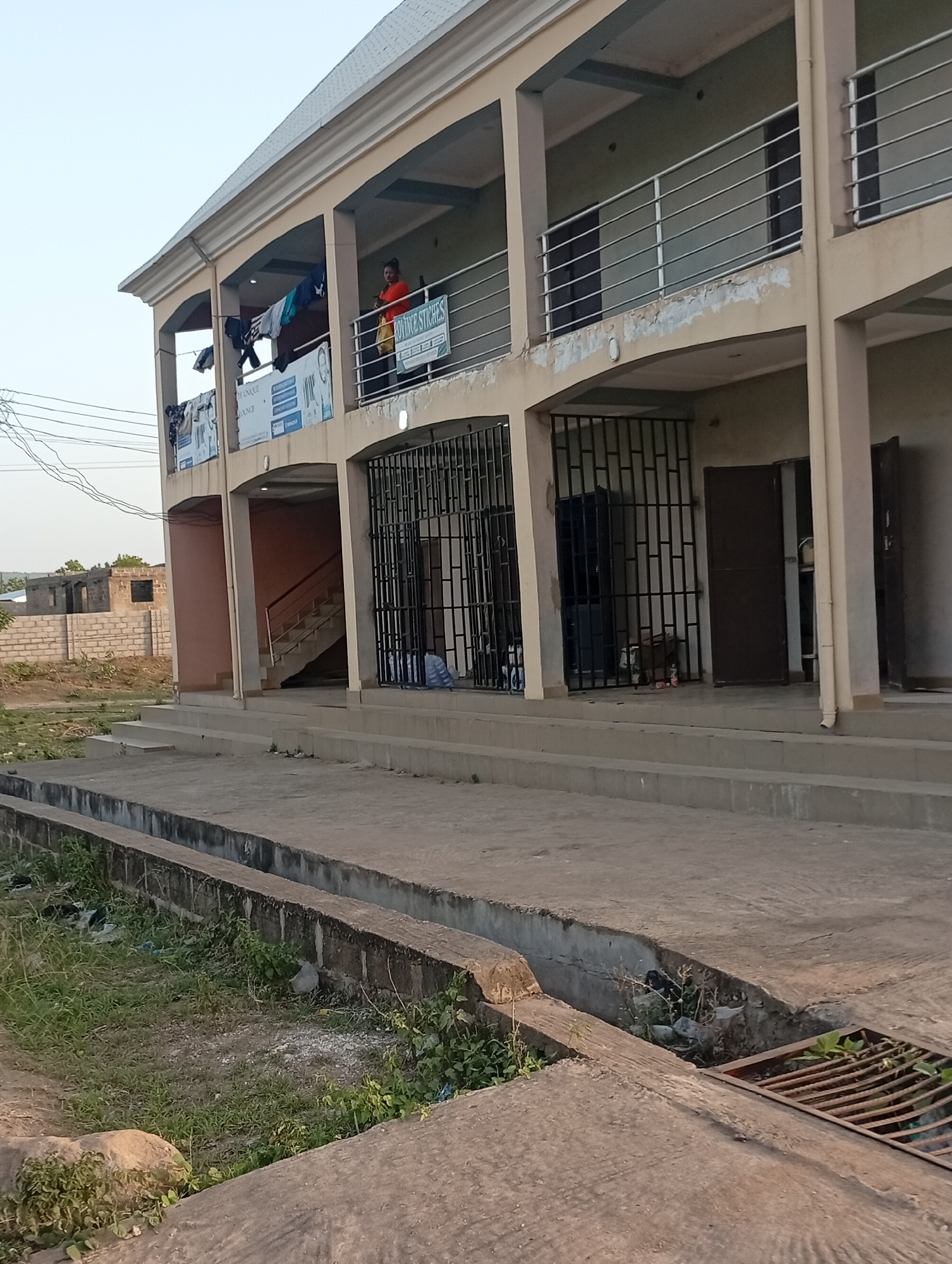 The scene depicts a two-story building in Adankolo, Nigeria. The building has open balconies with railings on the second floor. Clothing is hanging to dry on one balcony, and a woman in an orange shirt is standing on another. The ground floor has barred windows and open doorways. A concrete walkway with a drainage ditch runs along the front of the building. A partial view of a brick structure is visible in the background. Overhead power lines are also visible. The overall environment appears to be residential or commercial.