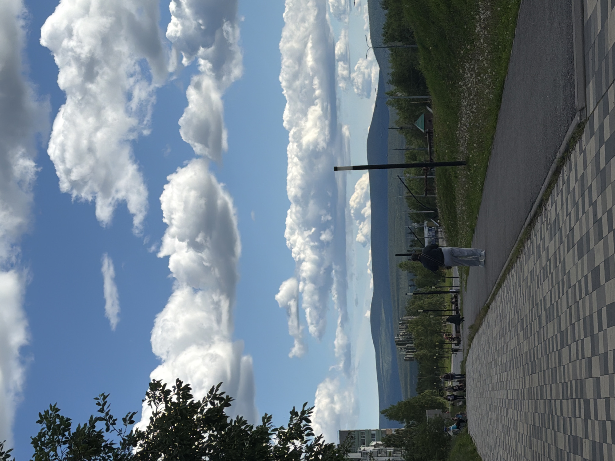 The image depicts a paved walkway in the city of Polarnie Zori, Russia, on a day with a bright blue sky dotted with fluffy white clouds. On the left side of the walkway, a person is walking away from the camera, wearing light-colored pants and a dark jacket. Further down the walkway, several other individuals are walking or standing, and some are seated on a bench. To the left of the walkway, a multi-story apartment building is visible, partially obscured by foliage. The background features a treeline with green foliage and distant, rounded mountains under the expansive sky. The walkway itself is constructed with rectangular paving stones arranged in a checkered pattern.