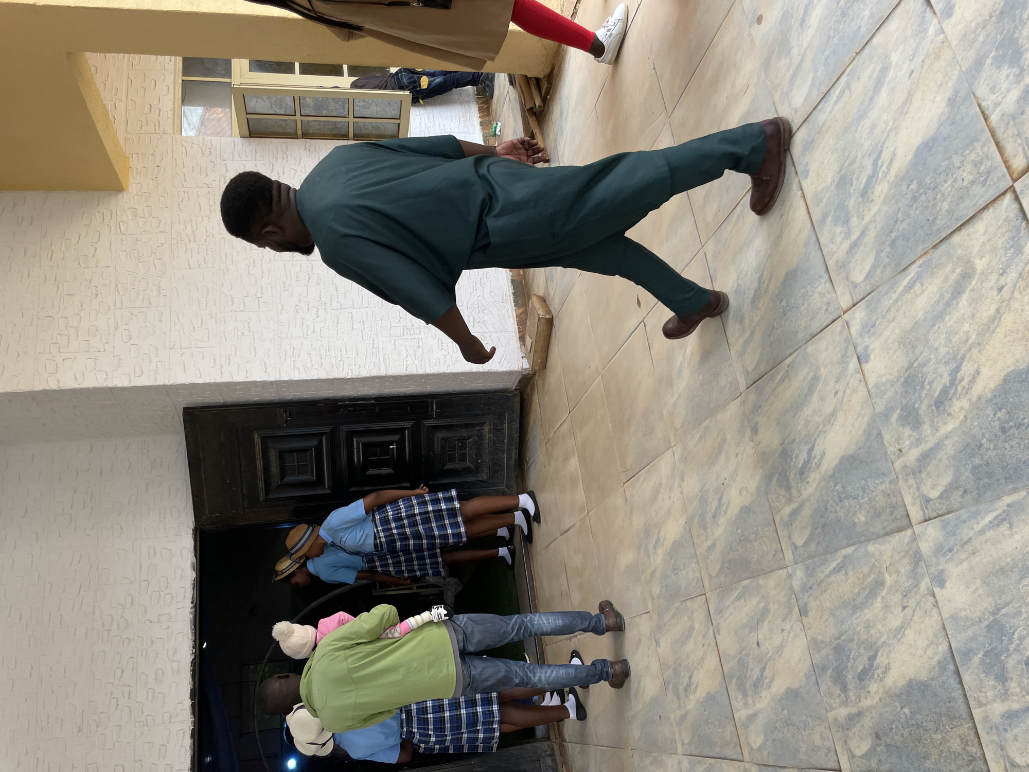 In Bar Gada, Nigeria, a man in a dark green traditional outfit walks across a tiled patio. He is moving away from a doorway where a group of young people in blue shirts and plaid skirts stand. One of the young people is holding a baby. A person in a light brown jacket and red leggings walks in the opposite direction, further into the background. The doorway is flanked by a white brick wall, and a yellow pillar supports an overhead structure.