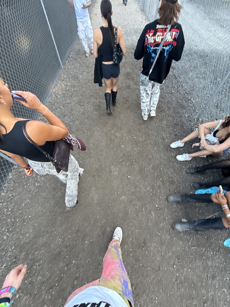 The image captures a view from above, looking down a dirt path in Indio, California. A chain-link fence runs along the left side. A group of individuals, presumably attendees of a music festival (possibly Coachella), is present.
People:
- Several people are walking along the path. They are dressed in various casual styles, including a black top with shorts and boots, a "Yu-Gi-Oh!" sweatshirt with patterned pants, and a person with tie-dye pants.
- A few individuals are sitting on the ground along the right side of the path.
Actions:
- People are walking along the path.
- Some people are sitting.
- One person is holding a phone.
Objects:
- Chain-link fence
- Dirt path
- Backpacks and purses
- A wristband.
Environment:
- The scene appears to be in an outdoor area with a dirt path, located in Indio, California. The presence of a chain-link fence and a dirt path suggests a temporary or event-specific setup.